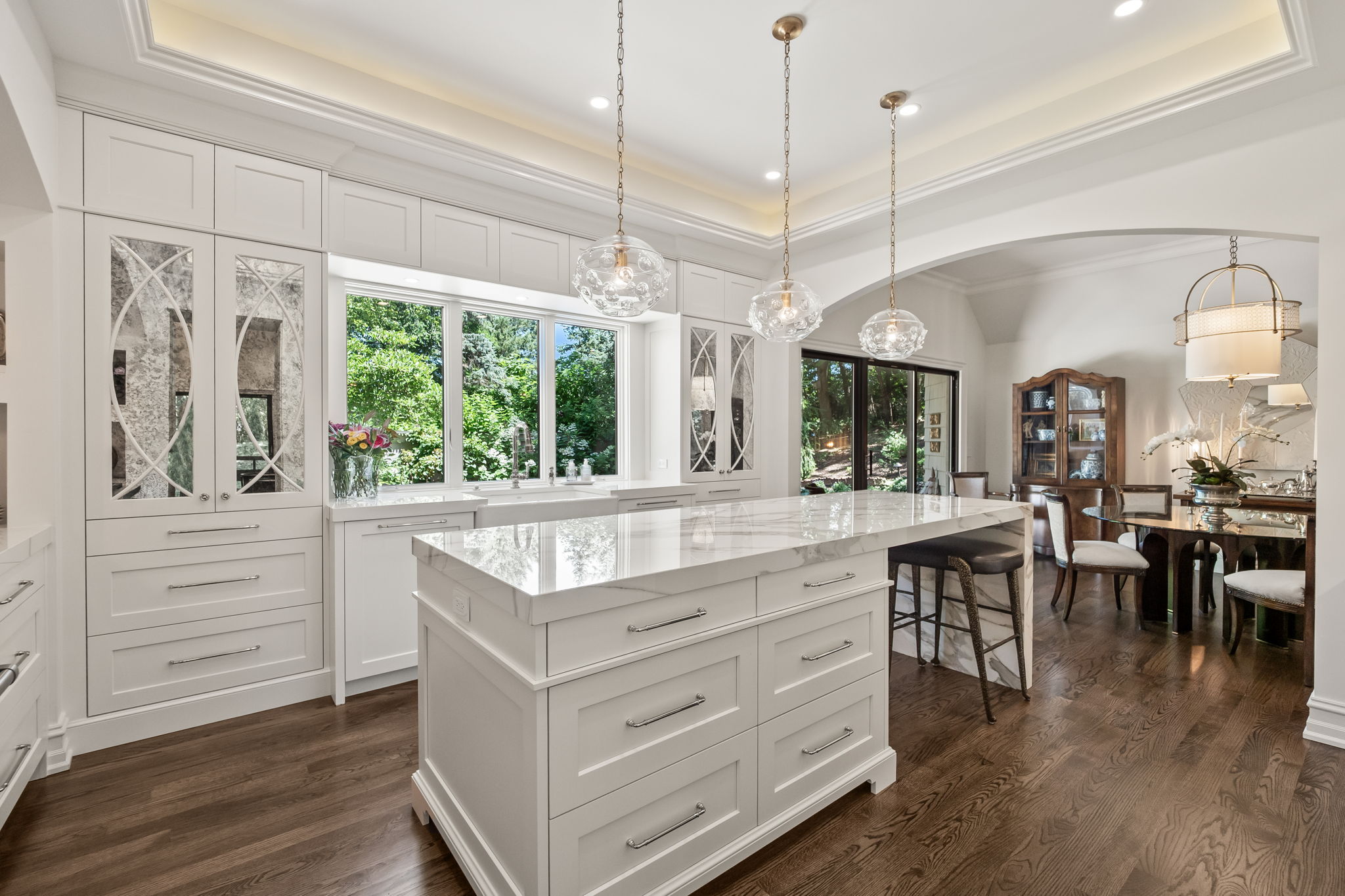 This is a bright and elegant kitchen featuring white cabinetry, a large marble-topped island with drawers, and pendant lighting. A large window provides natural light and a view of the greenery outside. The kitchen seamlessly transitions into a dining area, creating an open and inviting space.