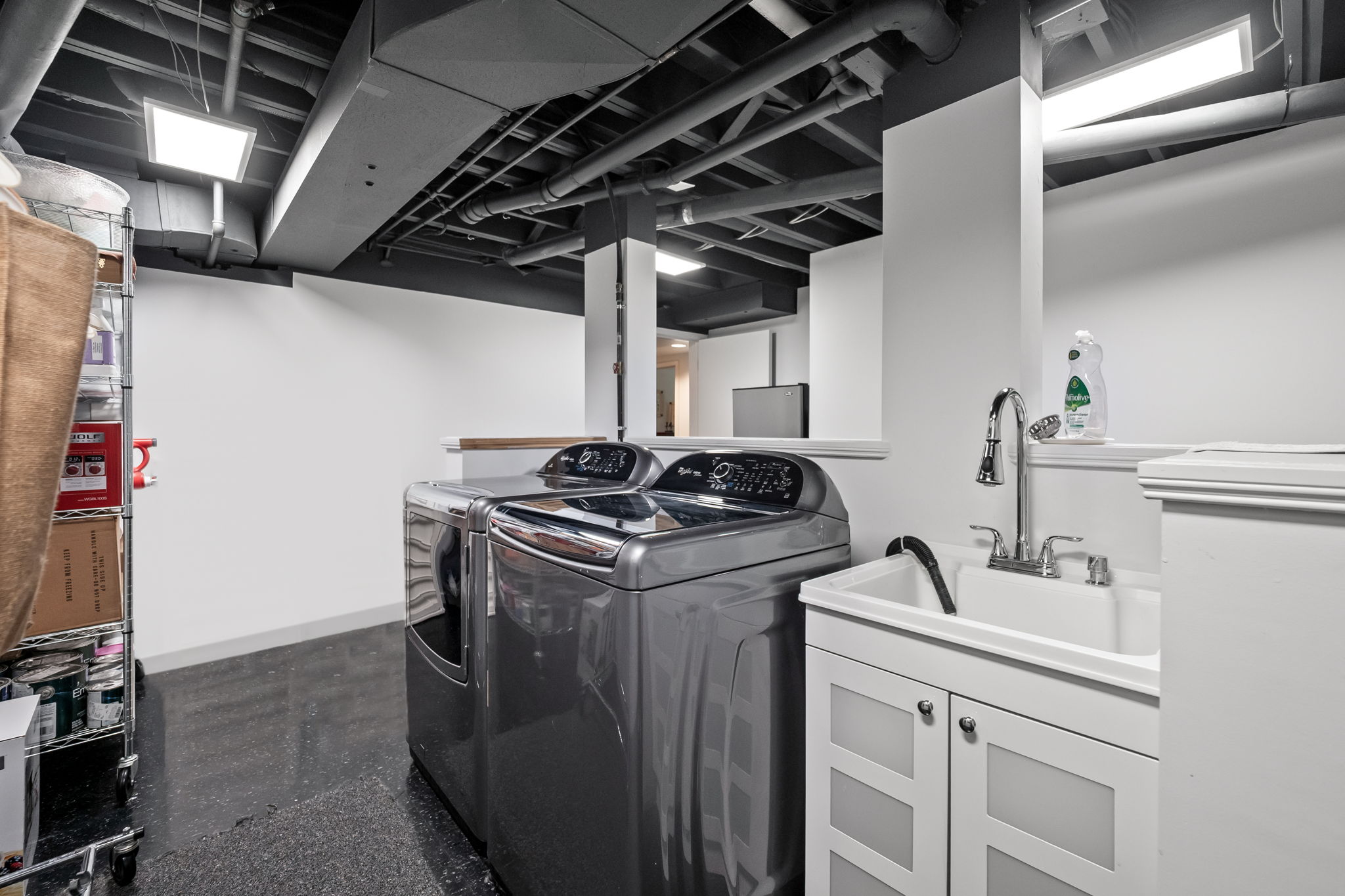 This is a well-organized laundry room featuring a modern washer and dryer set in a sleek gray finish. A utility sink with white cabinetry provides a practical space for handwashing and cleaning. The room has a clean and contemporary aesthetic with a dark floor and white walls, creating a functional and visually appealing space.