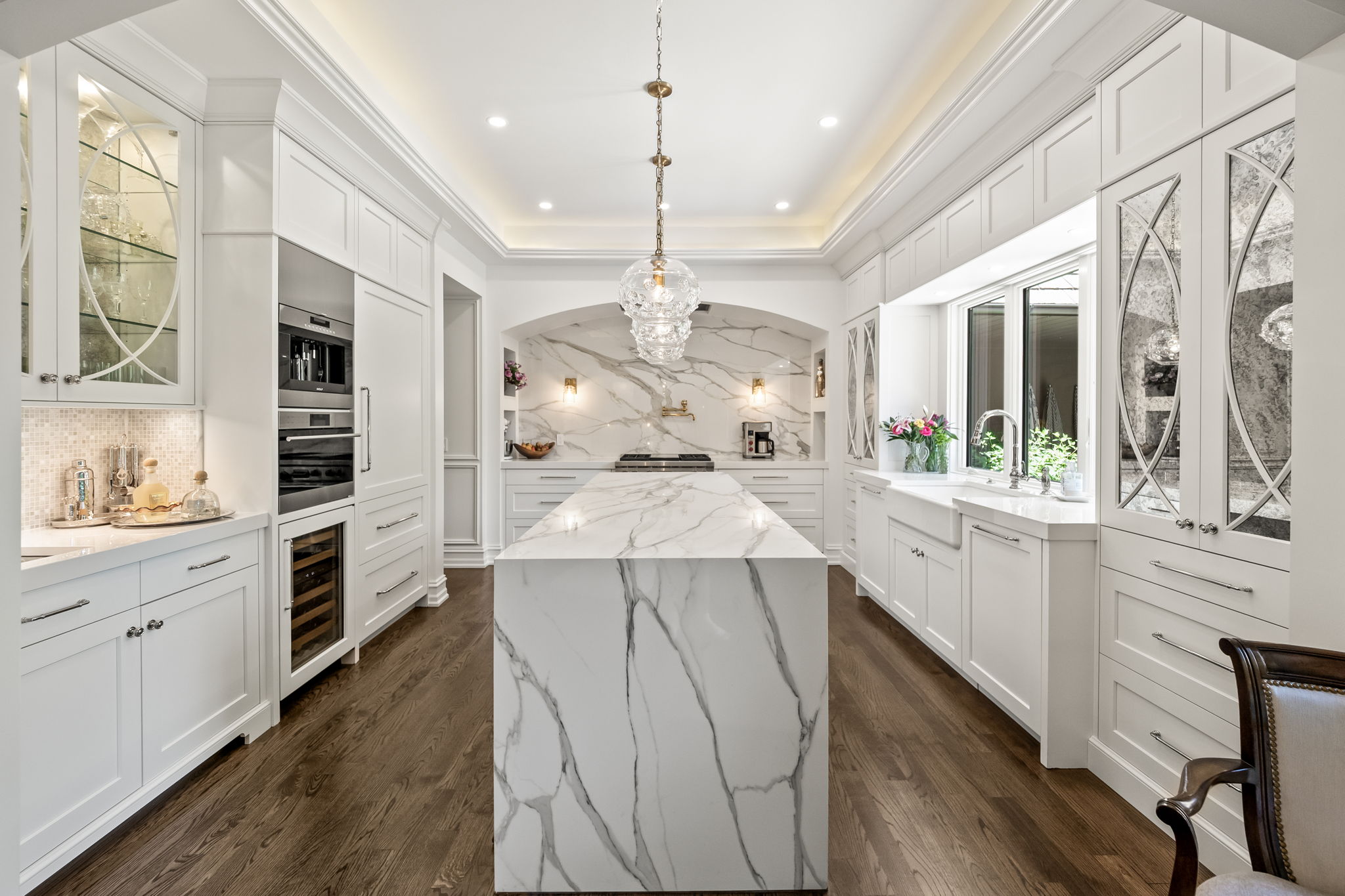 This is a bright and spacious kitchen featuring white cabinetry, marble countertops, and stainless steel appliances. A large island with a waterfall edge provides ample workspace, while a decorative chandelier adds a touch of elegance. The kitchen is well-lit with recessed lighting and natural light from a window above the sink, creating a clean and inviting atmosphere.