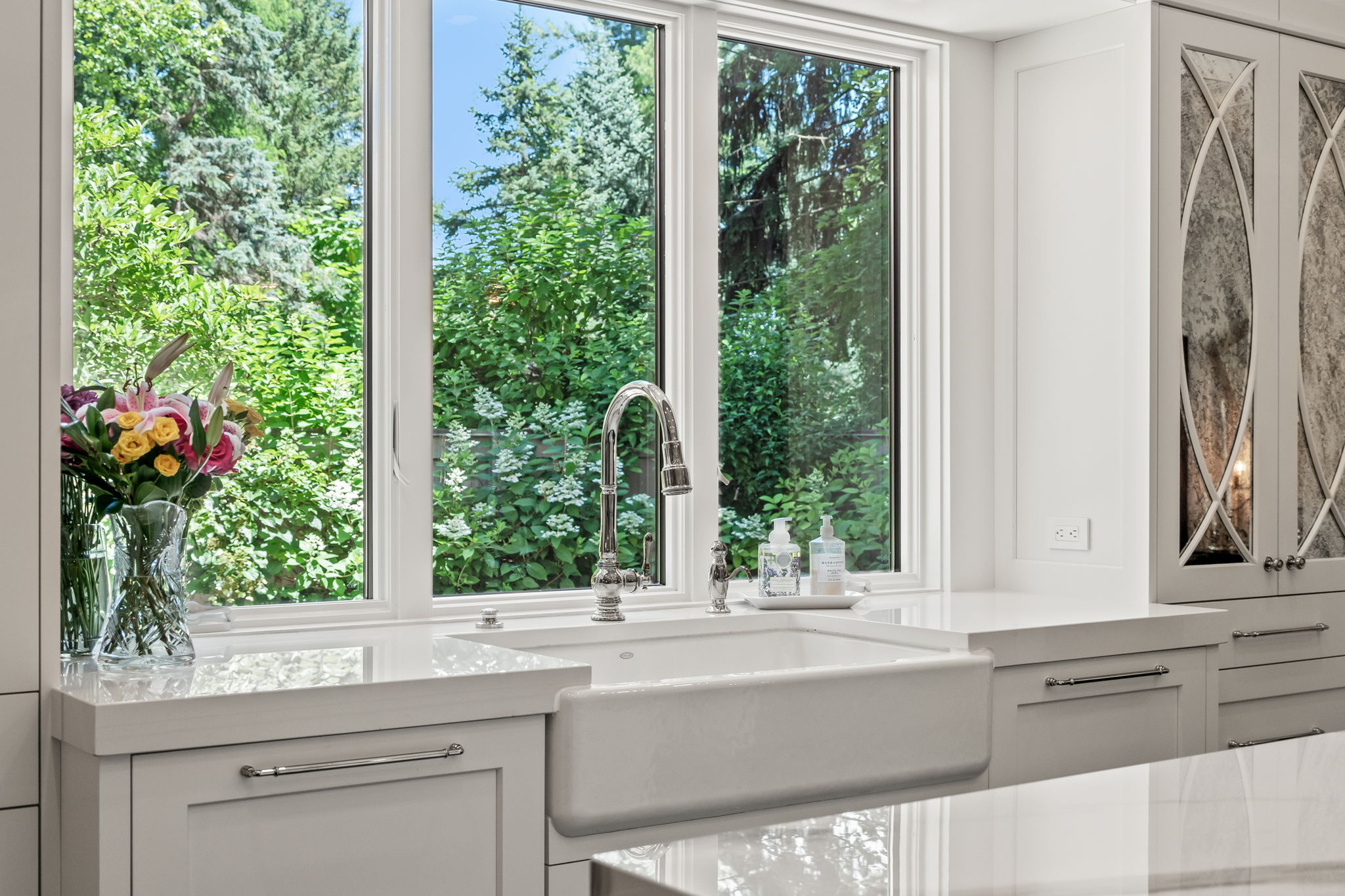 This is a bright and airy kitchen featuring a large farmhouse sink with a modern faucet, set beneath a window offering a view of lush greenery. The cabinetry is white with sleek hardware, complemented by light countertops and mirrored cabinet accents. The overall impression is clean, elegant, and inviting.