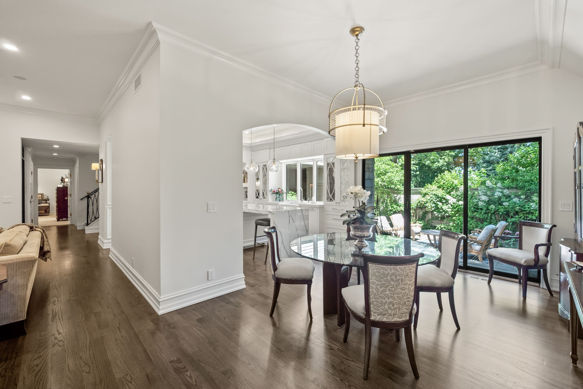 This is an interior shot of a dining room featuring a round glass-top table with four upholstered chairs. A modern chandelier hangs above the table, and a sliding glass door provides access to an outdoor patio area. The room has hardwood floors and white walls, creating a bright and elegant atmosphere.