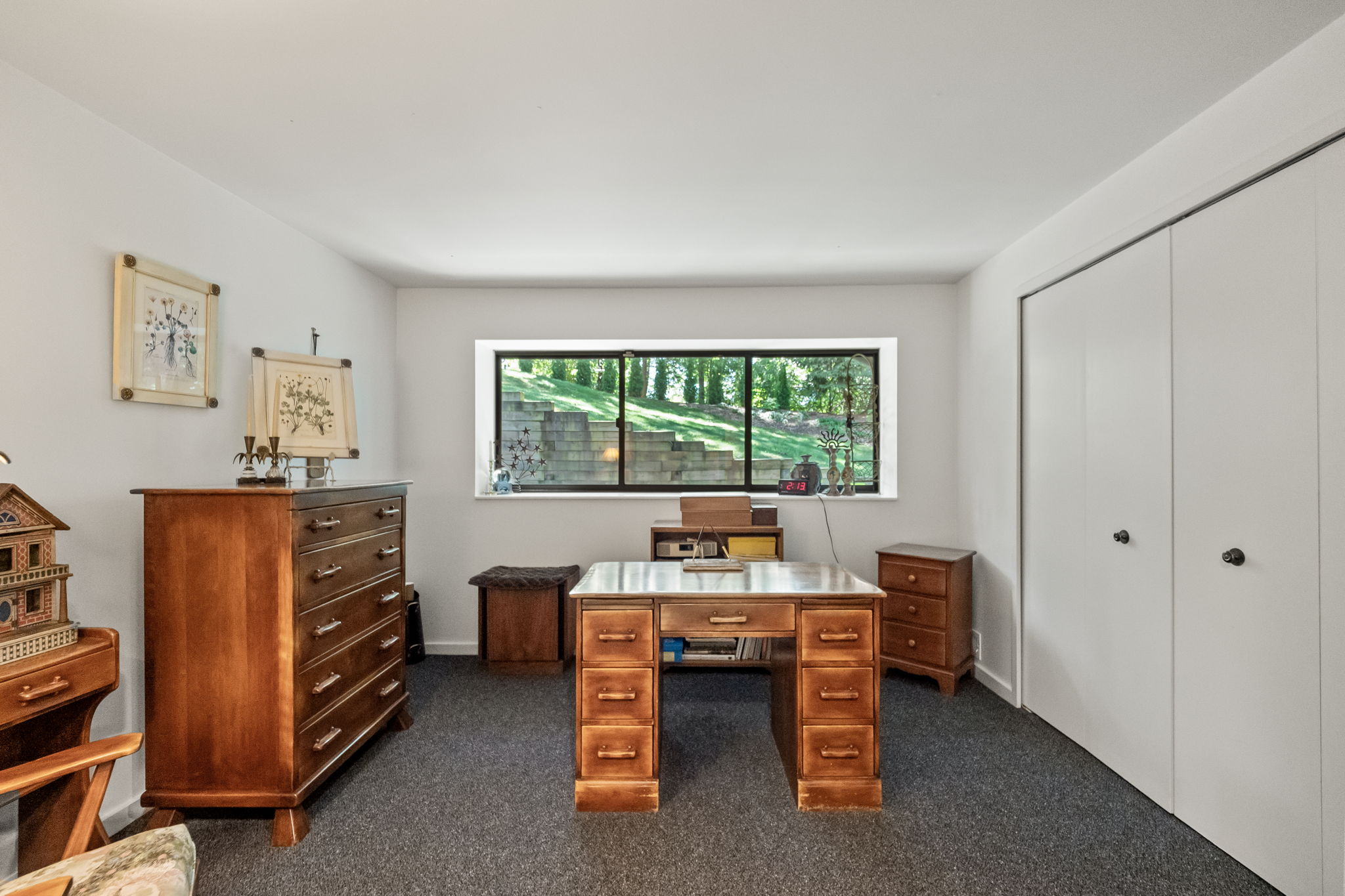 This is an interior shot of an office or study. The room features a wooden desk with multiple drawers, a matching dresser, and a small side table. A window provides natural light and a view of the outdoors, while a closet with white doors is visible on the right. The overall impression is a functional and organized workspace.