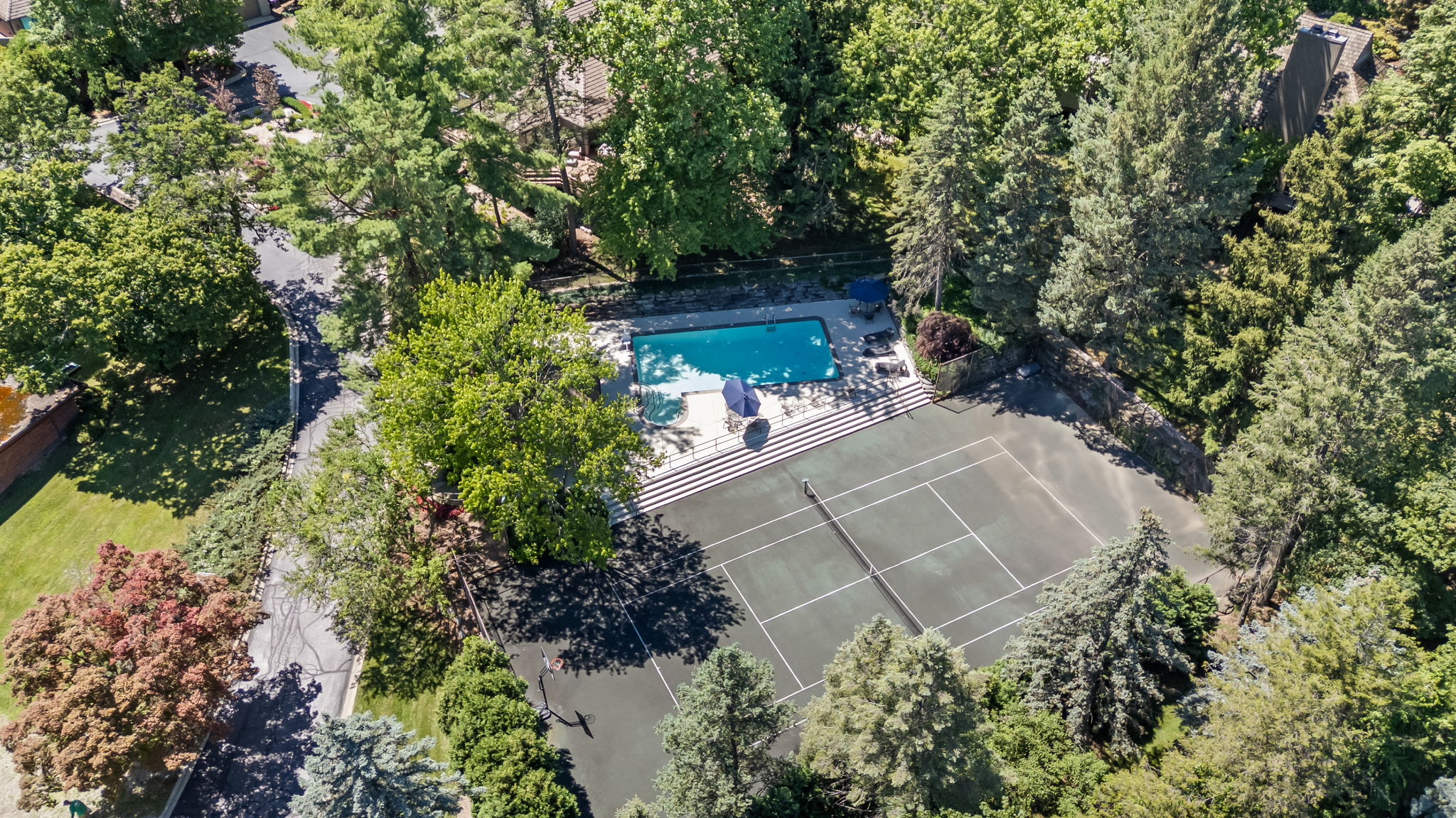 This aerial shot showcases a luxurious property featuring a tennis court and a swimming pool surrounded by lush greenery. The tennis court is well-maintained with clear lines, while the pool area includes lounge chairs and umbrellas for relaxation. The surrounding trees provide privacy and a sense of seclusion, enhancing the property's appeal.