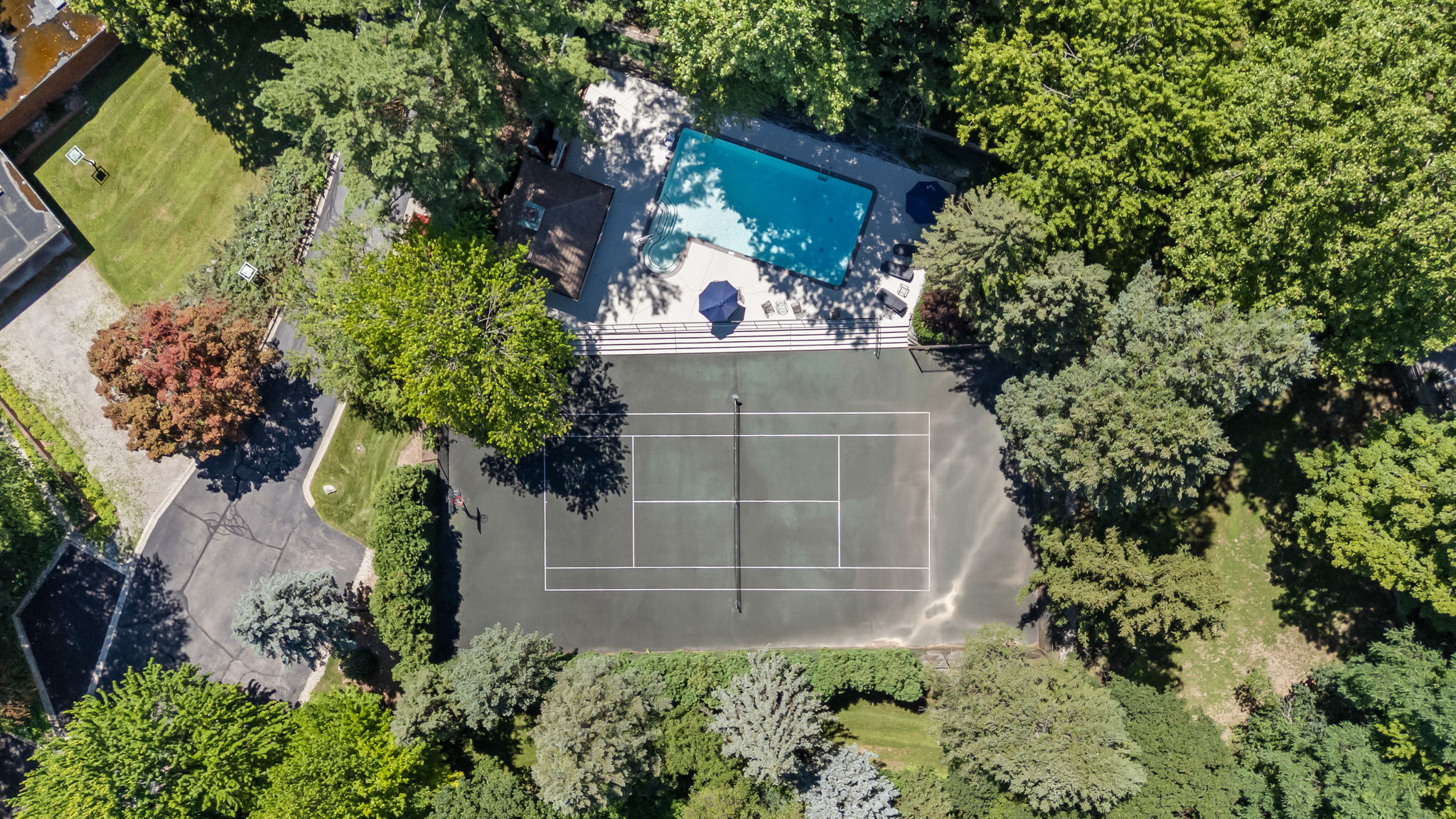 This aerial shot showcases a luxurious property featuring a tennis court, a swimming pool with a cabana, and lush landscaping. The tennis court is well-maintained with clear lines, while the pool area offers a relaxing retreat. Mature trees surround the property, providing privacy and a sense of seclusion.