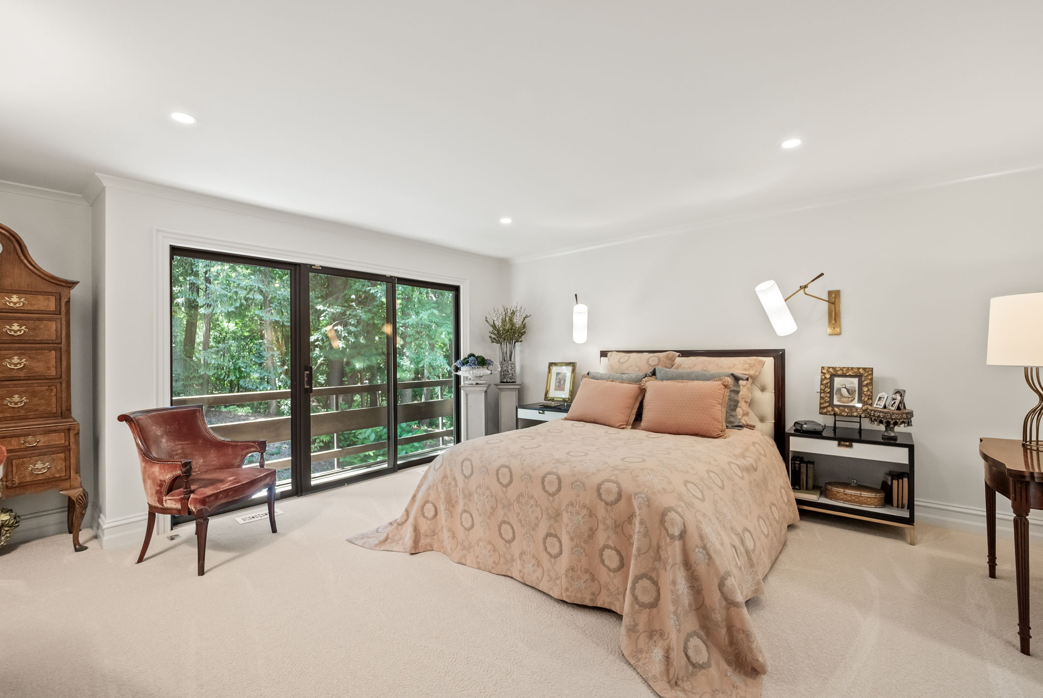 This is a well-lit primary bedroom featuring a large bed with a patterned peach-colored comforter and matching pillows. The room has a neutral color palette with white walls and light carpet. A sliding glass door leads to a balcony, providing natural light and a view of the surrounding greenery. There are two nightstands with lamps and decorative items on either side of the bed, and a vintage chair is placed near the sliding glass door.
