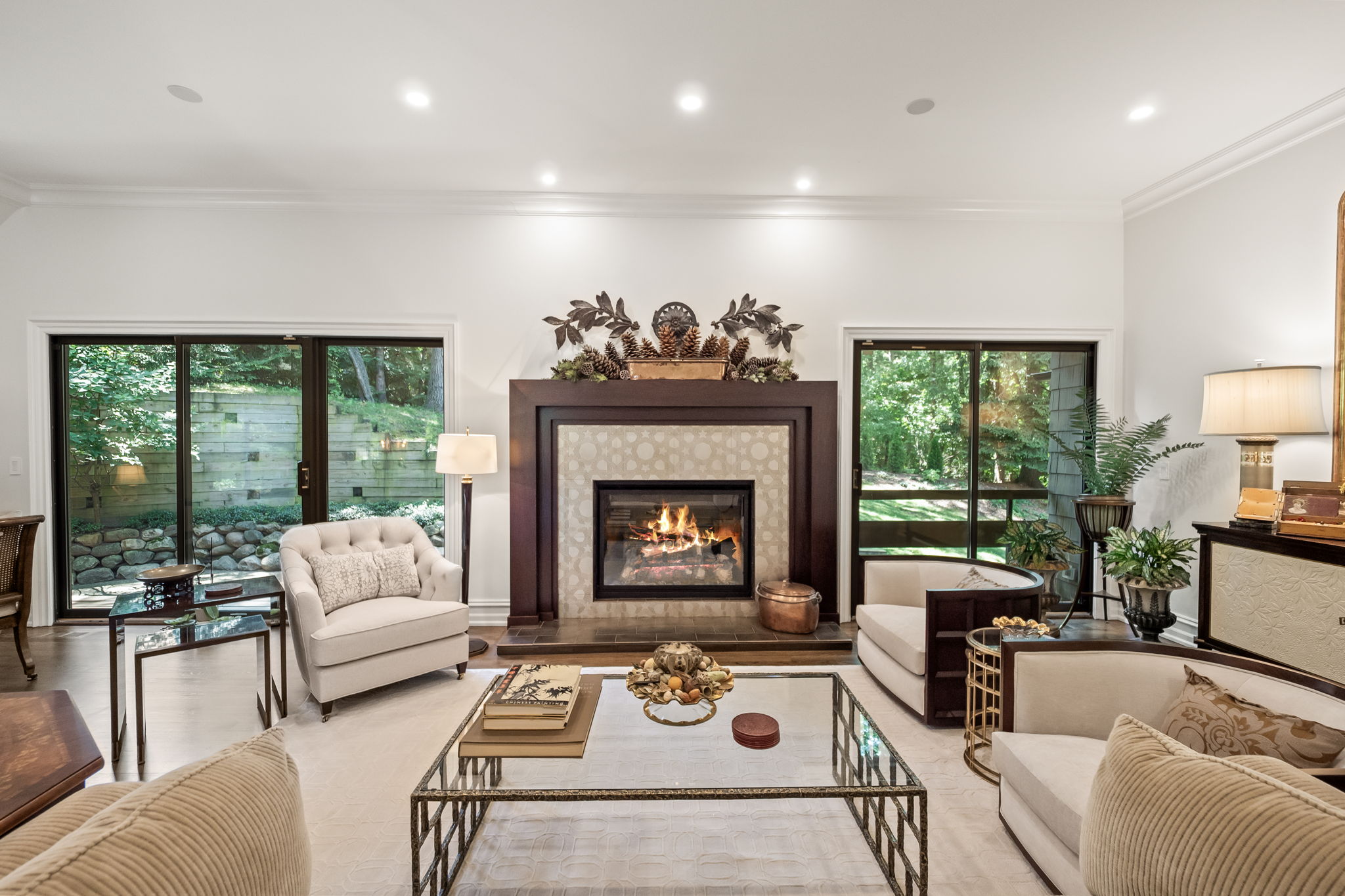 This is a well-lit living room featuring a fireplace as a focal point, flanked by large windows offering views of the outdoors. The room is furnished with comfortable seating, including armchairs and a sofa, arranged around a glass-topped coffee table. The decor is elegant and inviting, creating a warm and sophisticated atmosphere.