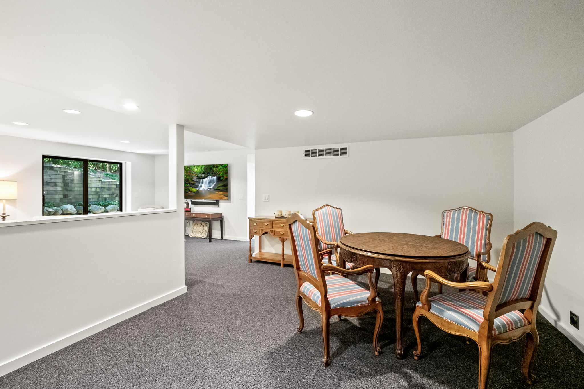 This is a well-lit basement area featuring a dining set with a round wooden table and four chairs with striped upholstery. The room has gray carpeting and white walls, with a partial wall separating it from another area where a television is mounted above a console table. The space feels clean and inviting, suitable for a recreation or dining area.