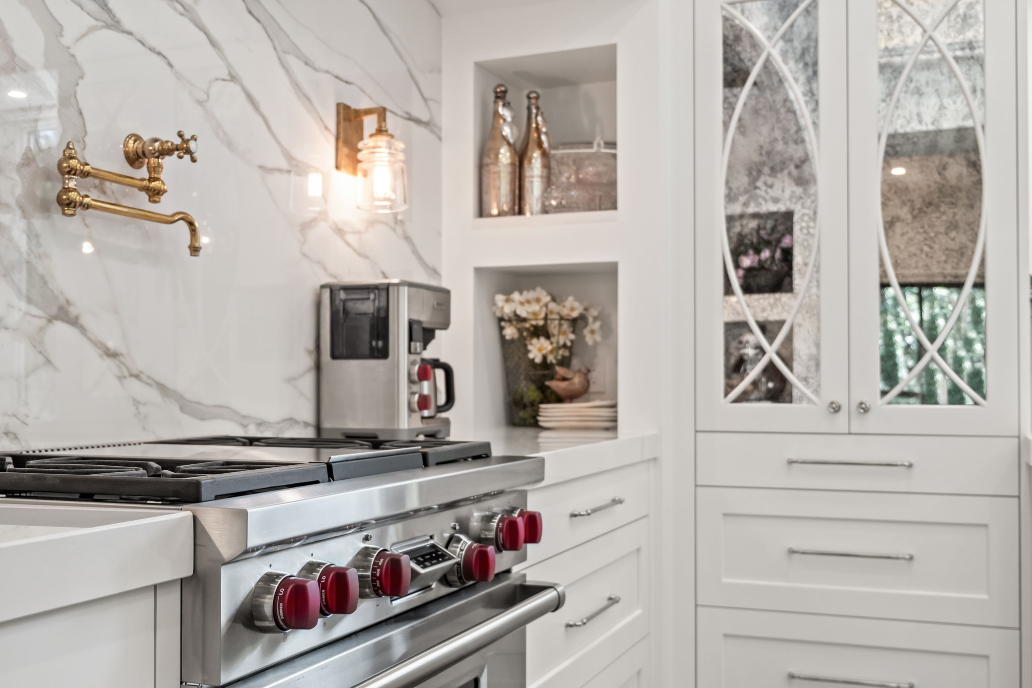 This is a detailed shot of a modern, high-end kitchen featuring a stainless steel range with red knobs, set against a backdrop of white marble backsplash and cabinetry. A pot filler faucet in gold adds a touch of luxury, while decorative niches and mirrored cabinets enhance the sophisticated aesthetic. The overall impression is one of elegance and functionality.