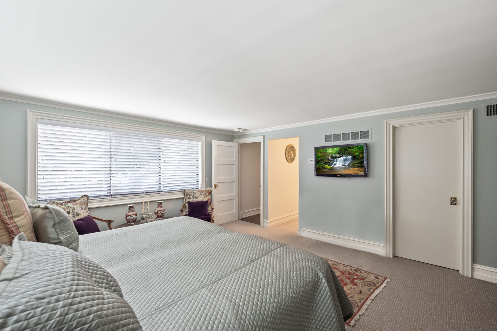 This is a primary bedroom featuring a large bed with a light blue quilted cover, two chairs near a window with blinds, and a wall-mounted television. The walls are painted a soft blue-gray, complemented by white trim and doors. The room has a calm and inviting atmosphere, perfect for relaxation.