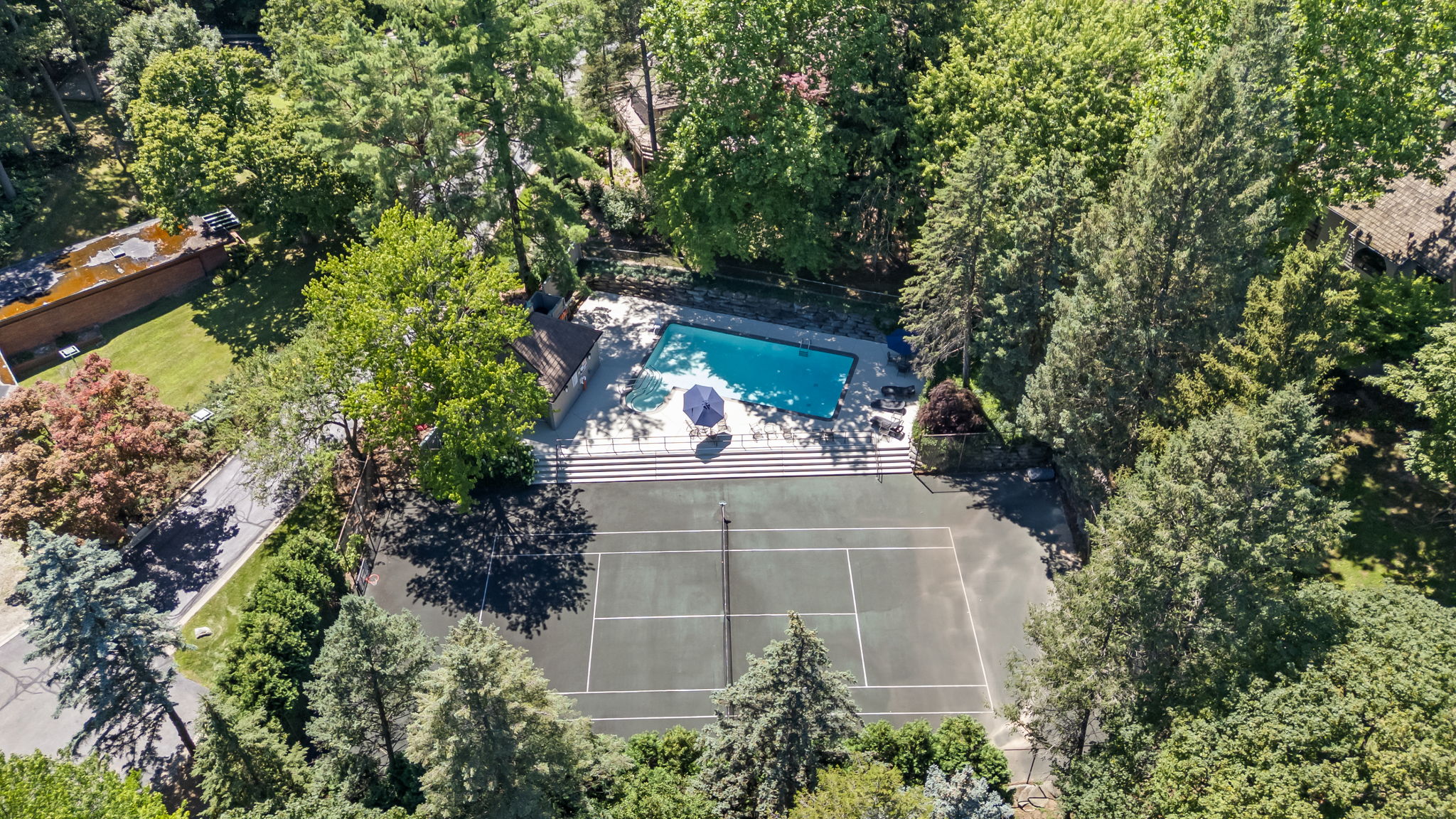 This aerial shot showcases a luxurious property featuring a tennis court and a swimming pool, both nestled among lush greenery. The tennis court is well-maintained with clear lines, while the pool area includes lounge chairs and an umbrella, suggesting a relaxing outdoor space. The surrounding trees provide privacy and a sense of seclusion, enhancing the property's appeal.