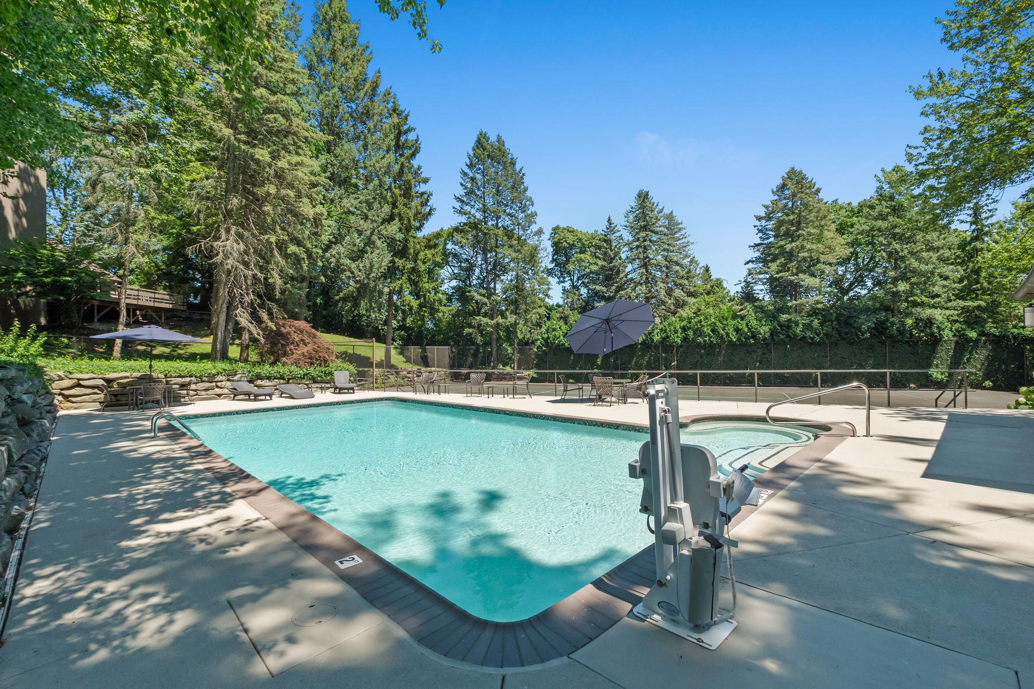This image showcases a pristine outdoor pool and spa area, surrounded by lush greenery and mature trees. The pool features clear, inviting water and is complemented by a spa with a safety railing. The surrounding patio area includes lounge chairs and tables with umbrellas, creating a relaxing and luxurious atmosphere.
