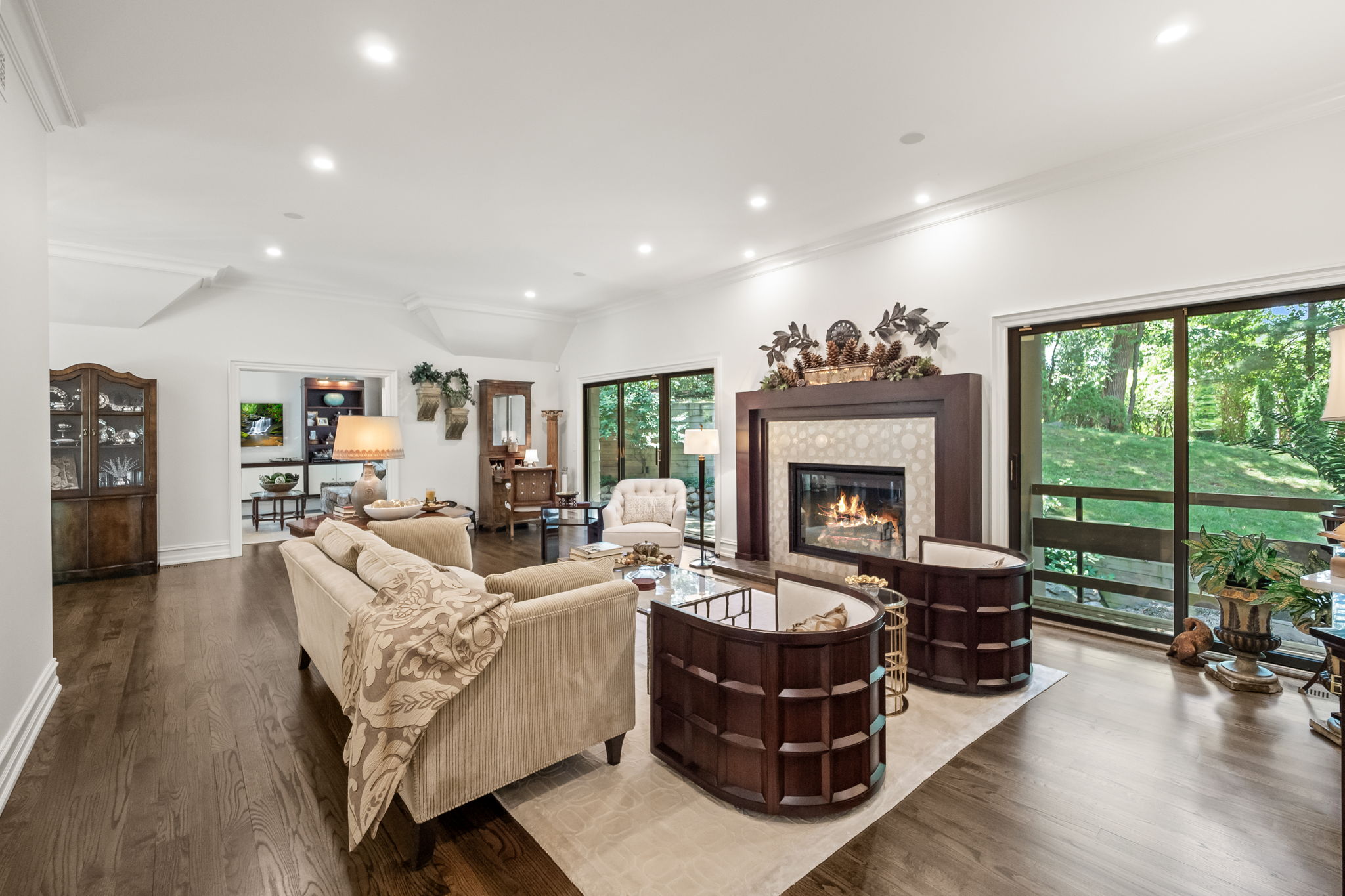 This is an interior shot of a living room featuring hardwood floors, a beige sofa with a patterned throw, and a fireplace with decorative elements above. The room is well-lit with recessed lighting and natural light coming through large sliding glass doors that lead to the outside. The overall impression is one of warmth and elegance.