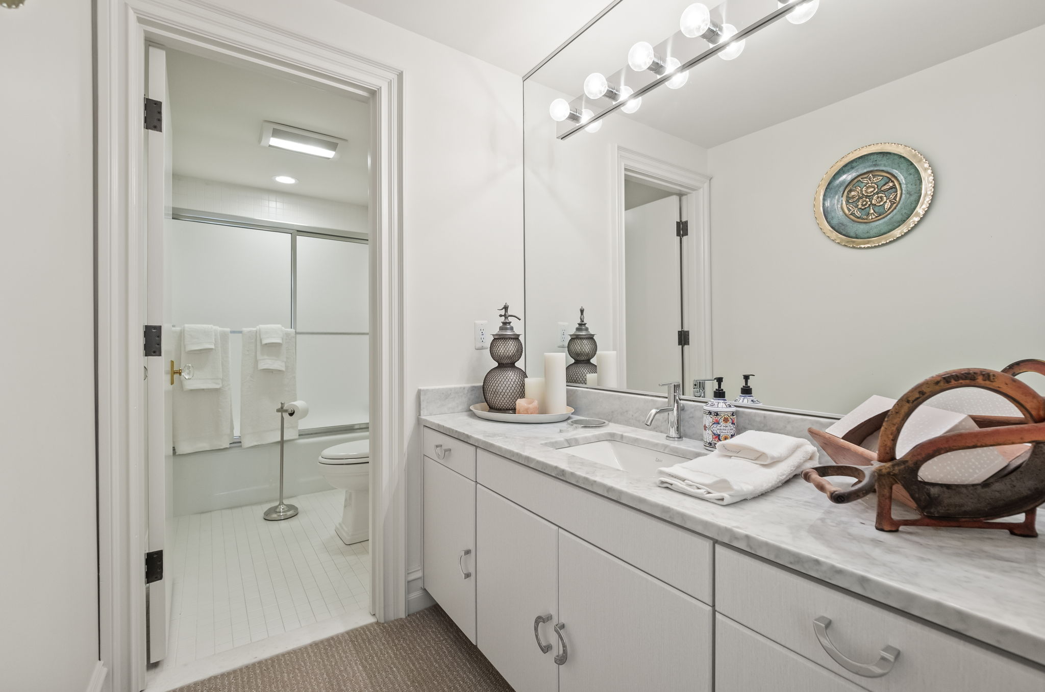 This is a well-lit bathroom featuring a white vanity with a marble countertop, a rectangular sink, and chrome fixtures. A large mirror with vanity lighting hangs above the sink, and decorative items add a touch of style. The bathroom includes a toilet and a shower/tub combination with a sliding glass door, all contributing to a clean and functional space.