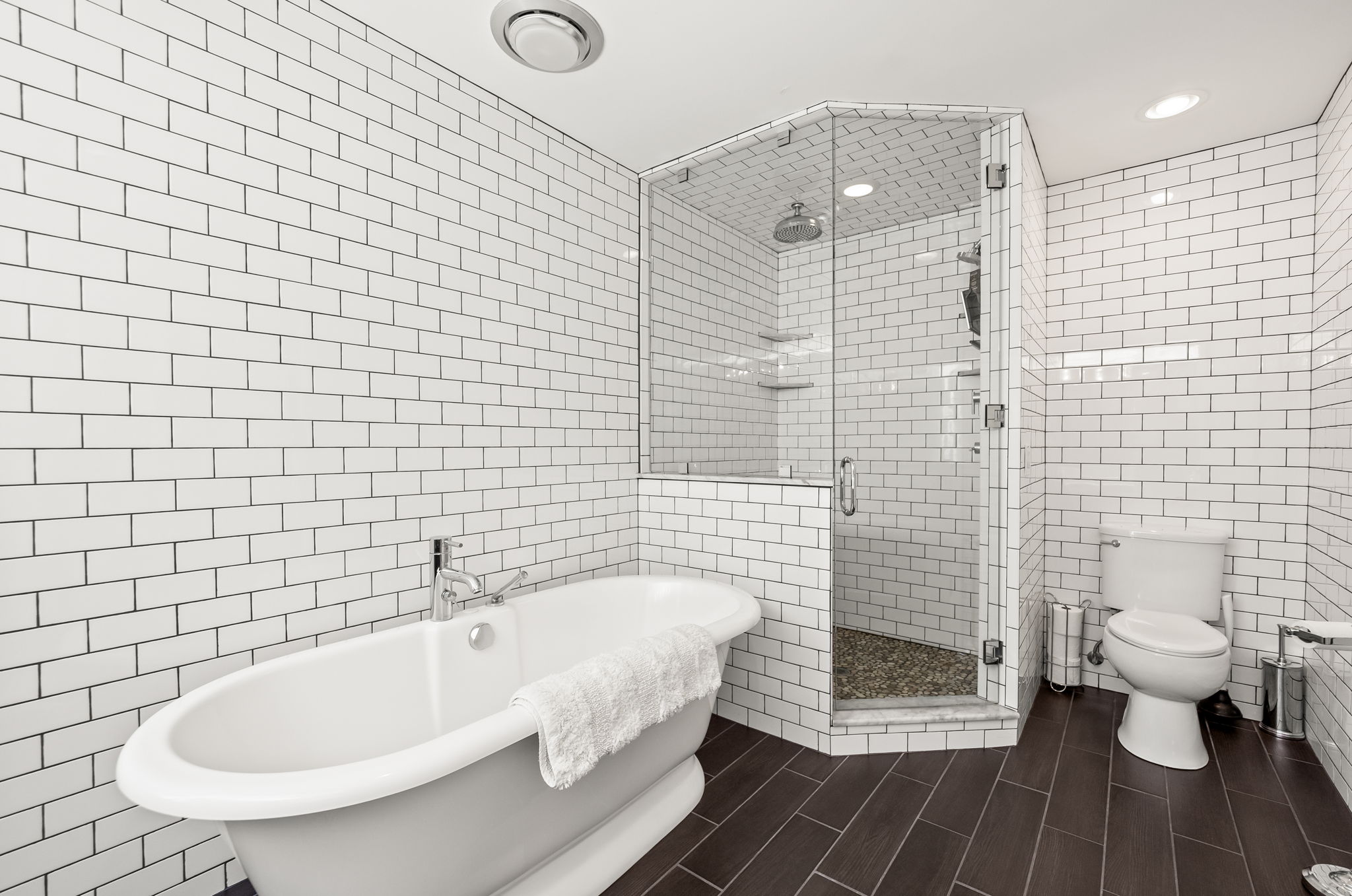 This is a well-lit primary bathroom featuring white subway tile walls and dark wood-look flooring. A freestanding bathtub with chrome fixtures sits to the left, while a glass-enclosed shower is positioned in the corner. A white toilet is located on the right side of the frame, completing the classic and clean aesthetic of the space.