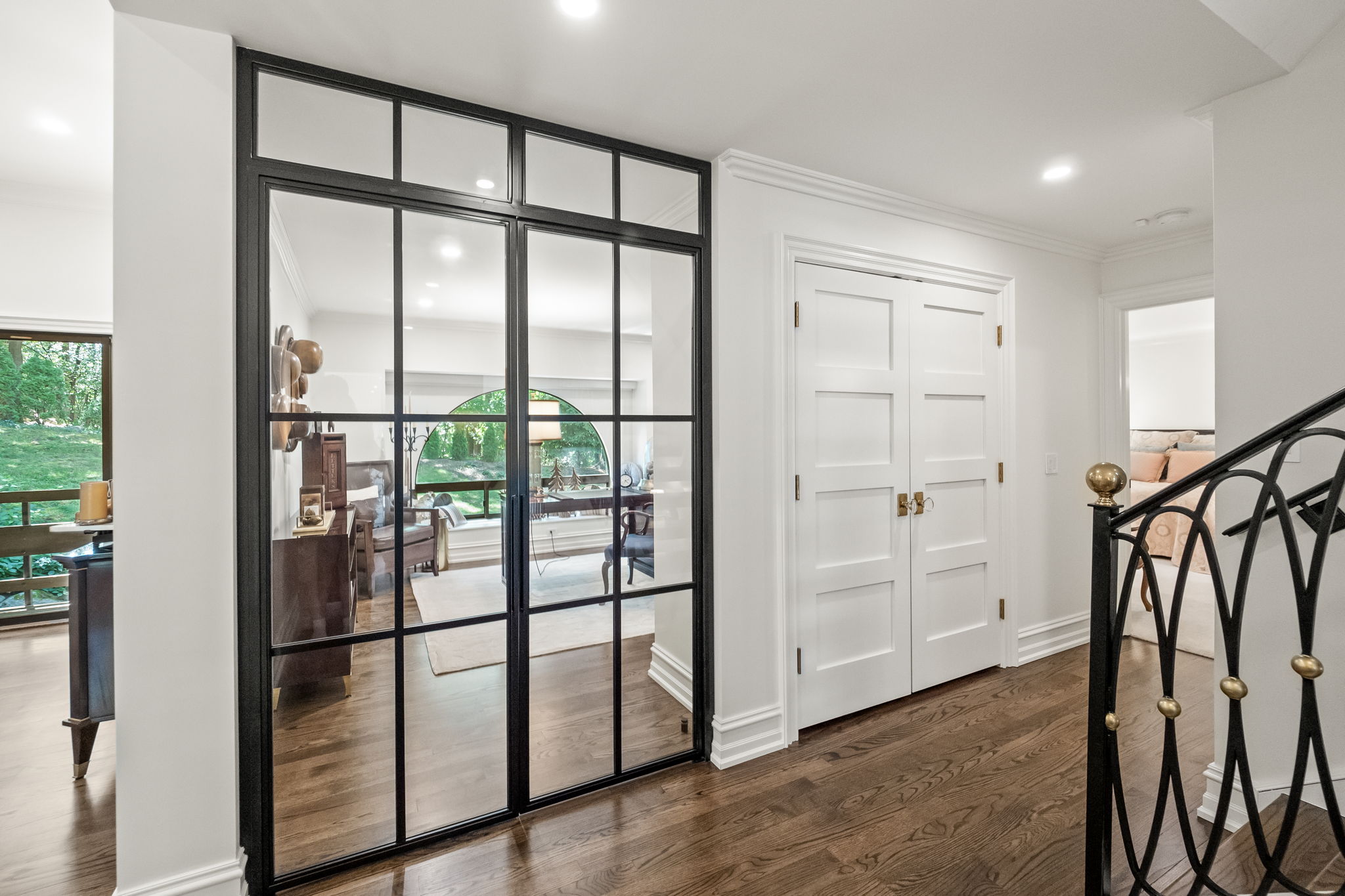 This interior shot showcases a bright hallway with dark hardwood flooring and white walls. A black-framed glass partition separates the hallway from another room, while a set of white paneled doors is visible on the right. A black wrought-iron staircase with gold accents adds a touch of elegance to the space.