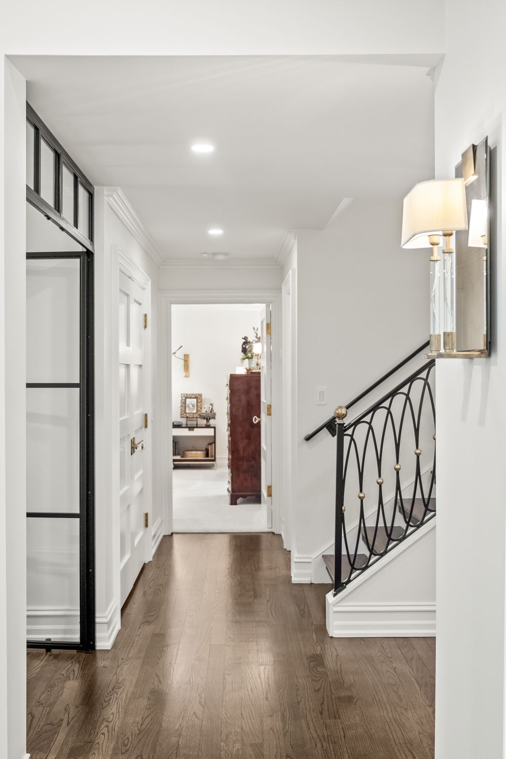 This interior shot showcases a well-lit hallway with dark hardwood floors and white walls, leading to an open doorway revealing a glimpse of another room. A black metal and gold accented staircase is visible on the right, complemented by a decorative wall sconce. The space exudes a sophisticated and clean aesthetic, highlighting the home's attention to detail.