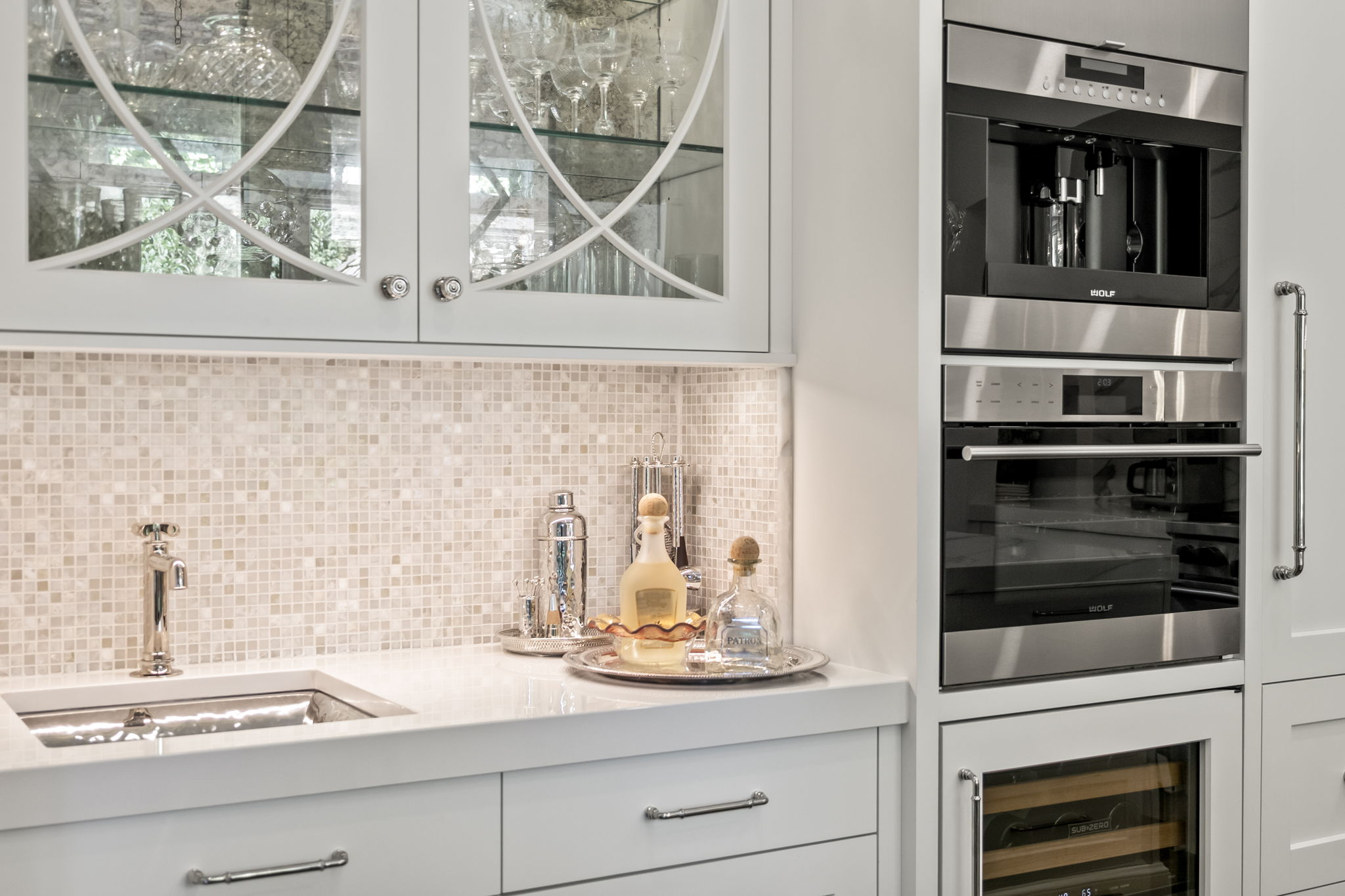 This image showcases a well-appointed kitchen area featuring white cabinetry, a mosaic tile backsplash, and stainless steel appliances, including a Wolf coffee maker and oven. A stylish sink and faucet are visible, along with decorative bottles and a cocktail shaker on the countertop. The overall impression is one of luxury and sophistication.