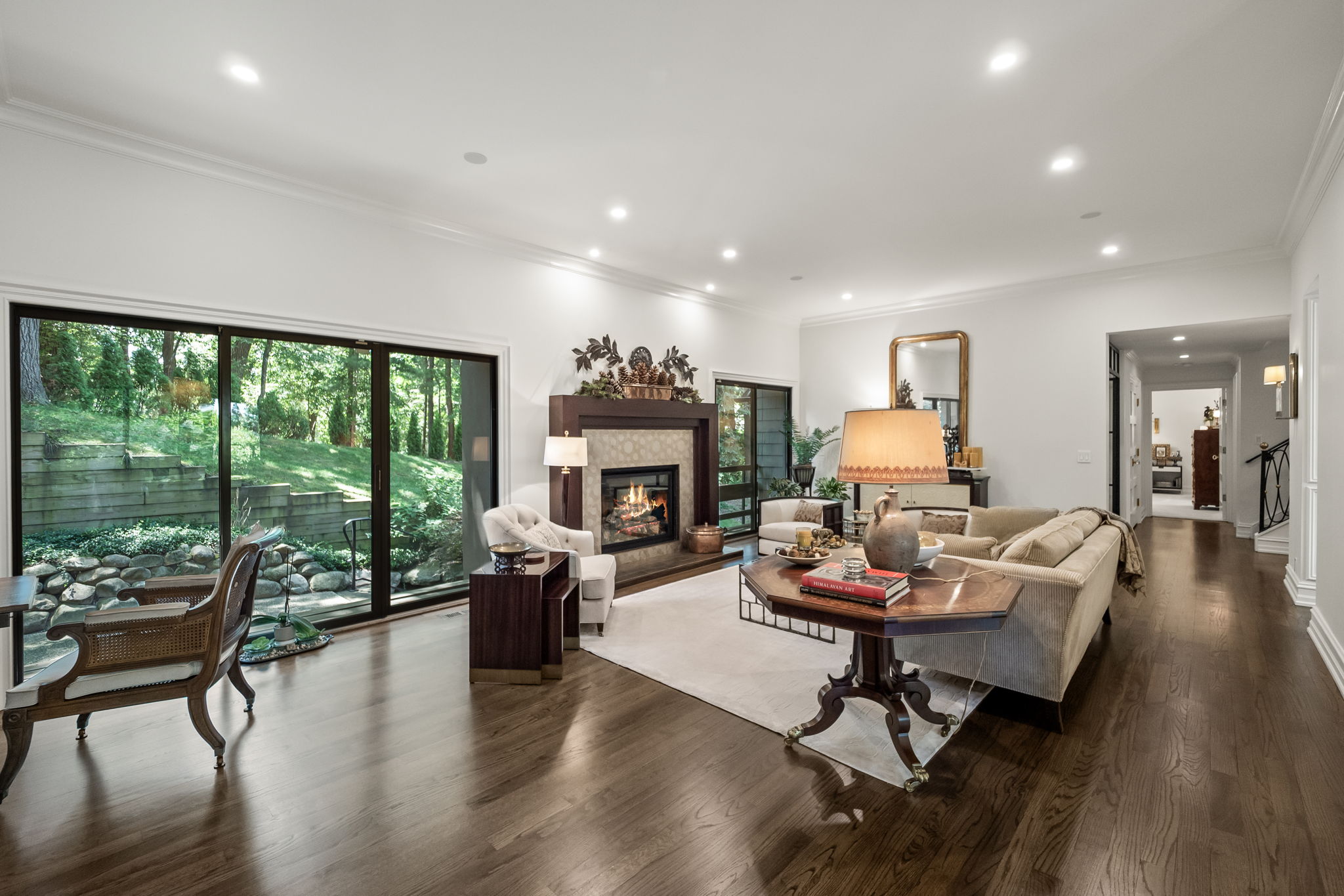 This is an interior shot of a well-lit living room featuring hardwood floors, white walls, and recessed lighting. A fireplace with decorative mantle serves as a focal point, complemented by comfortable seating arrangements and a large window offering a view of the outdoor landscape. The room exudes a sense of warmth and sophistication, ideal for relaxation and entertaining.