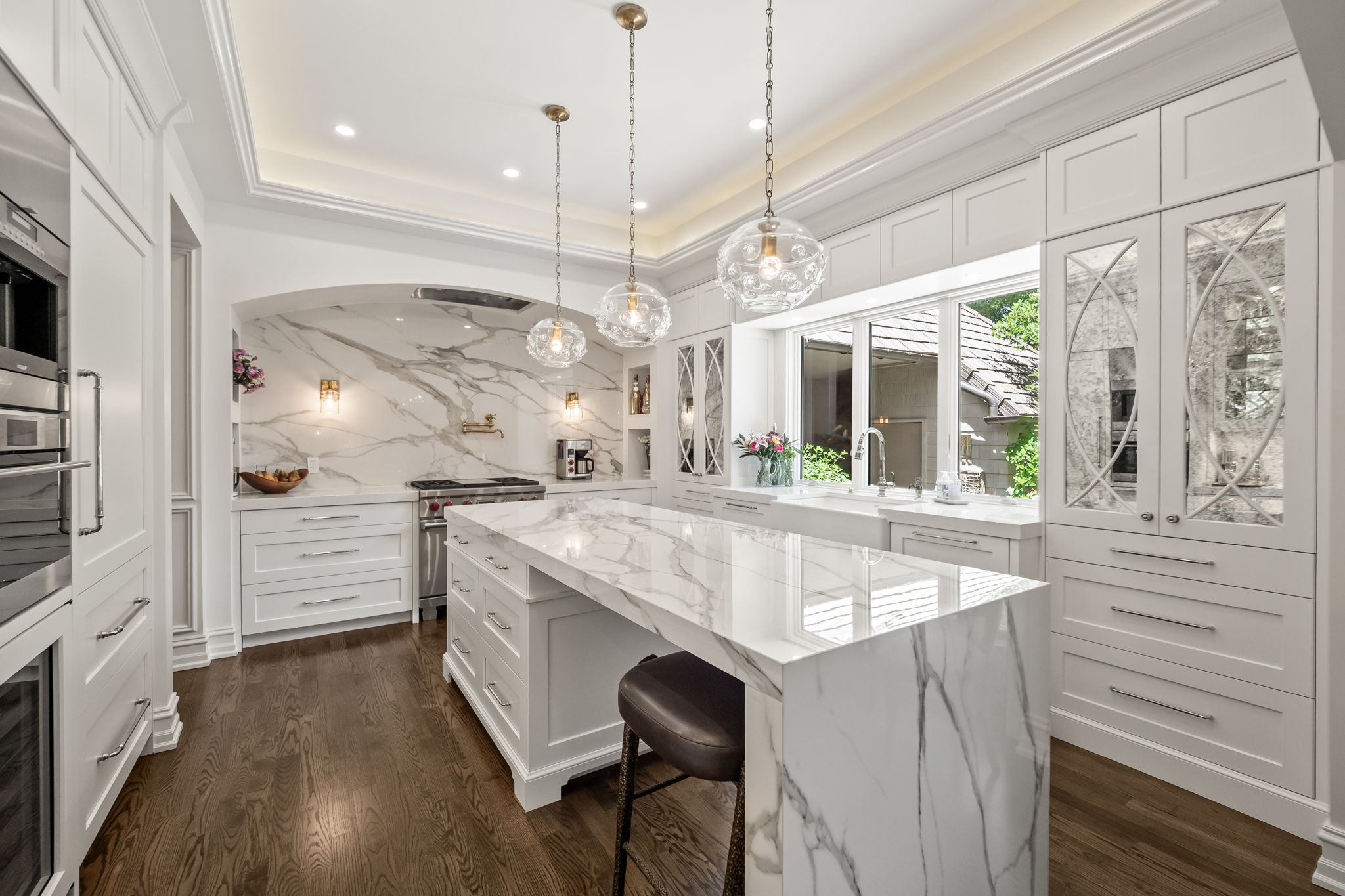 This is a bright and luxurious kitchen featuring white cabinetry, marble countertops, and stainless steel appliances. The kitchen island is a focal point, complemented by pendant lighting and a window that provides natural light. The overall impression is one of elegance and high-end design, perfect for a modern home.