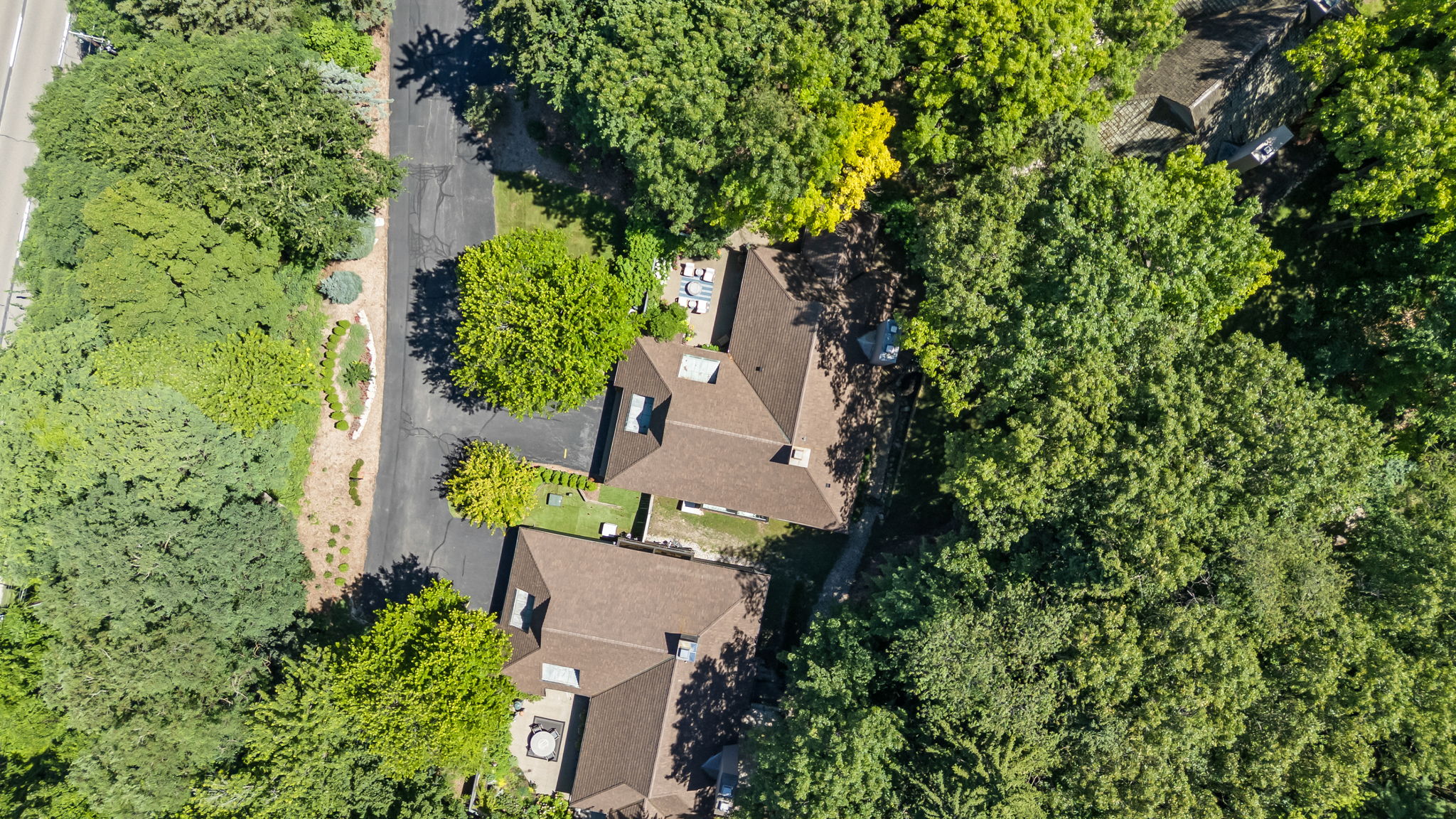 This aerial shot showcases a cluster of houses nestled among lush green trees, offering a sense of privacy and seclusion. The houses feature brown roofs and are surrounded by dense foliage, creating a serene and natural environment. A road runs alongside the properties, providing access while maintaining a tranquil atmosphere.