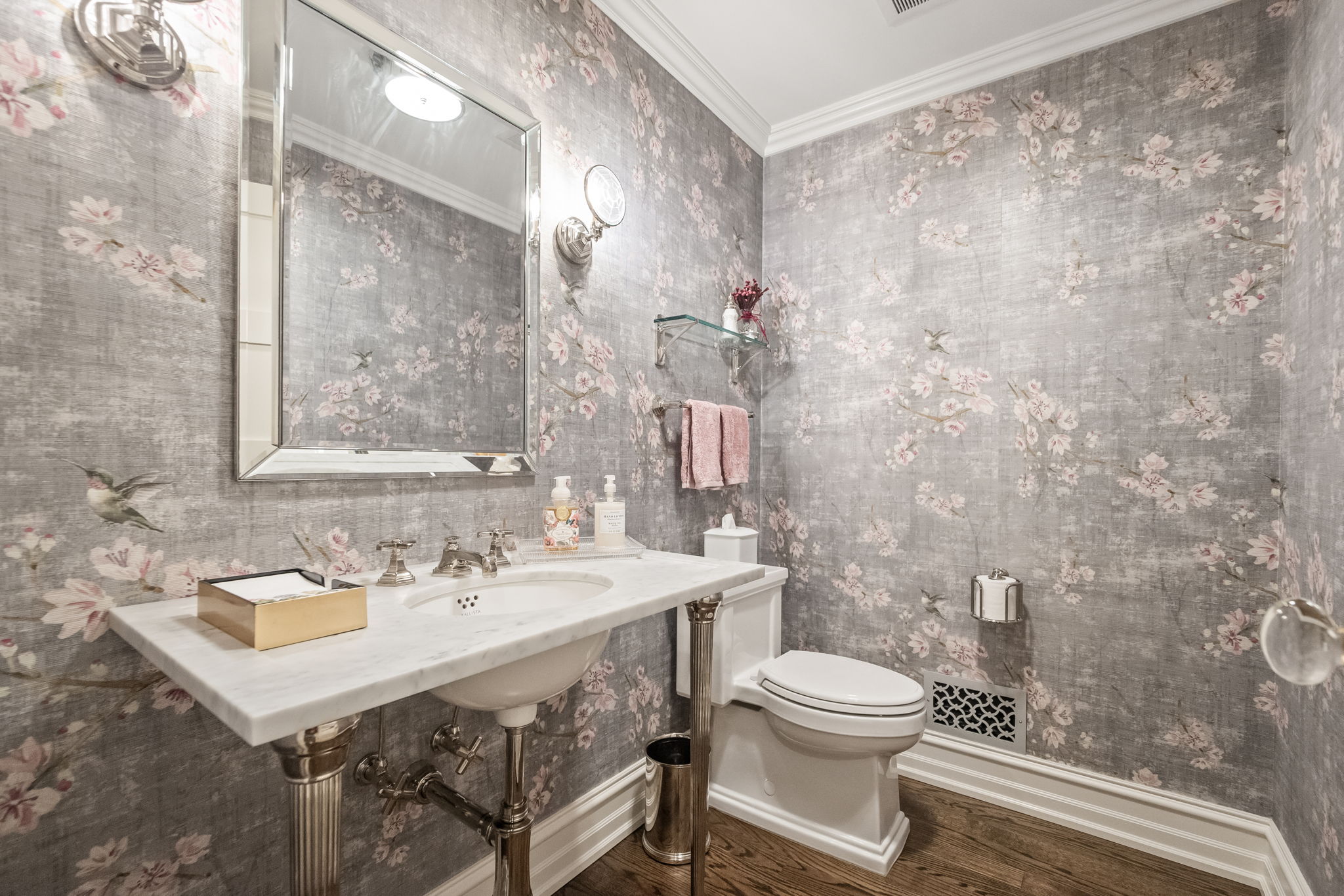 This is a stylish guest bathroom featuring a pedestal sink with a marble countertop and silver fixtures. The walls are adorned with a floral wallpaper, creating a sophisticated and elegant ambiance. A toilet and a small glass shelf with decorative items complete the space, which is illuminated by sconces and a mirror with integrated lighting.