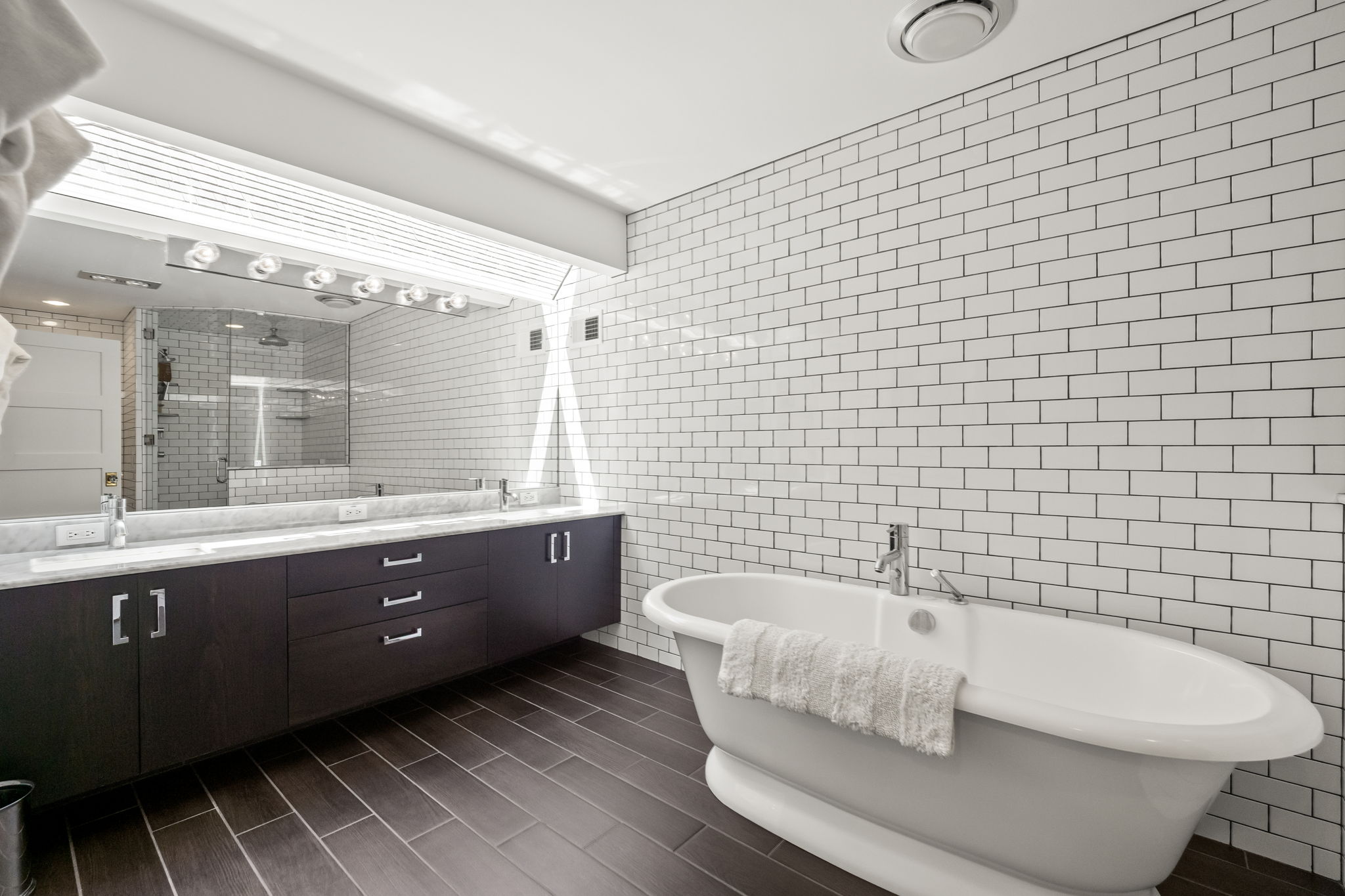 This is a well-lit primary bathroom featuring a freestanding white bathtub with a towel draped over the side, a dark wood vanity with double sinks and a marble countertop, and a large mirror with vanity lighting. The walls are covered in white subway tile with dark grout, and the floor is dark wood. The overall impression is clean, modern, and luxurious.