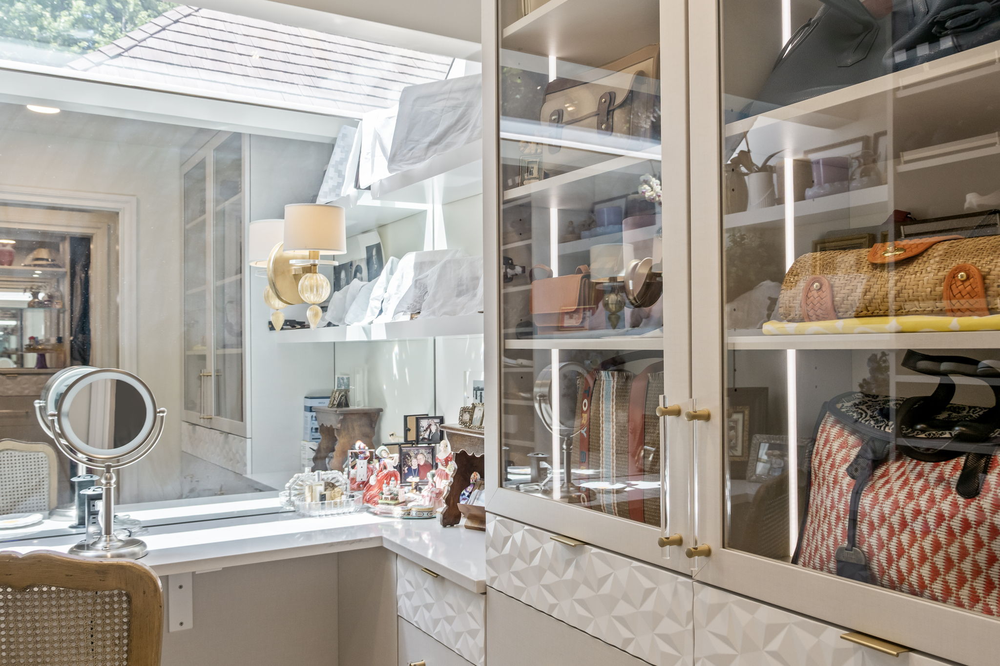 This image showcases a luxurious walk-in closet with custom cabinetry and ample storage space. The closet features glass-fronted cabinets displaying handbags and accessories, complemented by drawers with geometric detailing and gold hardware. A vanity area with a lighted mirror adds a touch of elegance, creating a well-organized and stylish dressing space.