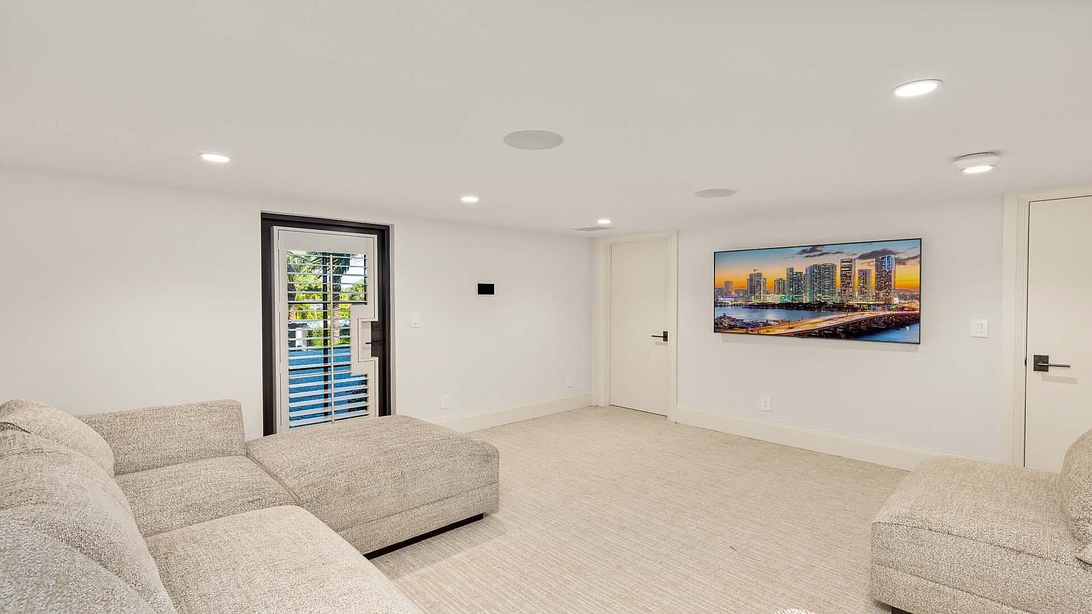 The image showcases a modern living room with a neutral color palette. A large sectional sofa and armchair provide ample seating, while a flat-screen TV mounted on the wall serves as a focal point. A door with shutters leads to the outside, and recessed lighting illuminates the space, creating a bright and inviting atmosphere.