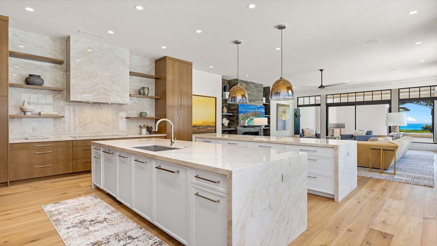 This is a bright and modern kitchen featuring two large islands with white cabinetry and light-colored countertops. The kitchen includes stainless steel appliances, open shelving, and pendant lighting. Large windows offer a view of the ocean, creating a luxurious and inviting atmosphere.