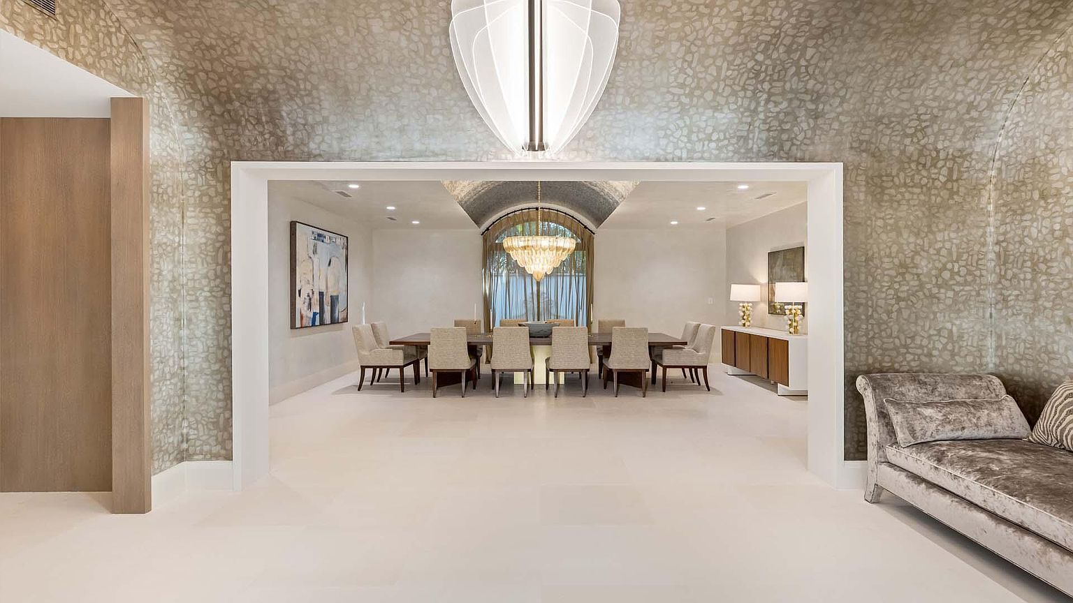 This image showcases a luxurious dining room, viewed through a wide doorway, featuring a long wooden table surrounded by upholstered chairs. A grand chandelier hangs above the table, and the room is bathed in natural light from a large arched window. The walls are adorned with textured wallpaper, and a modern chaise lounge sits to the side, adding to the room's elegant and inviting atmosphere.