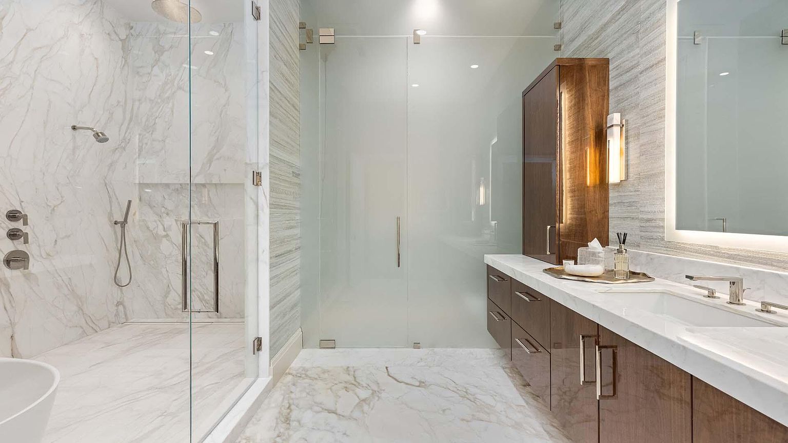 This is a luxurious primary bathroom featuring marble flooring and walls, a glass-enclosed shower with modern fixtures, and a vanity with a marble countertop and dark wood cabinetry. The space is well-lit with recessed lighting and a decorative wall sconce, creating a spa-like atmosphere. The overall impression is clean, elegant, and high-end.