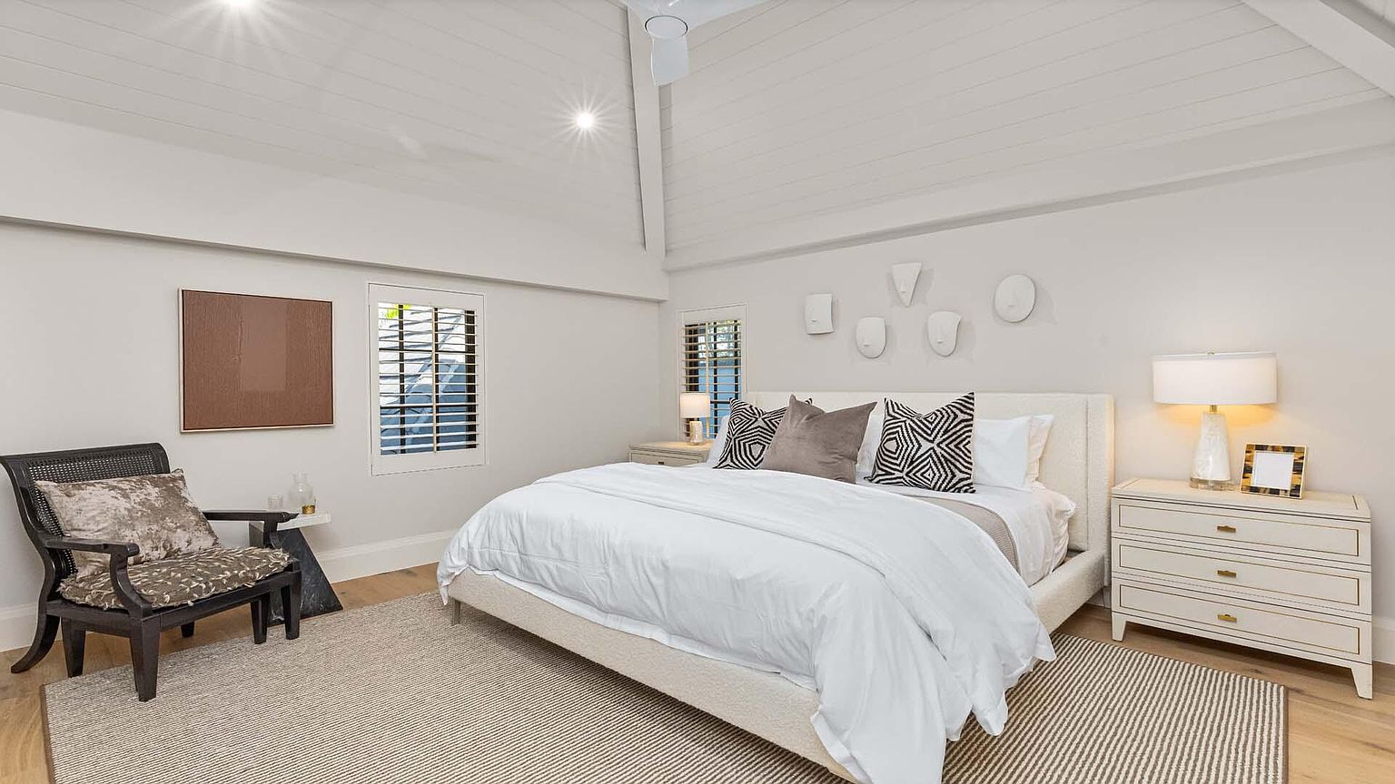 This is a bright and airy primary bedroom featuring a neutral color palette. The room includes a large bed with white linens, decorative pillows, and a textured headboard. A nightstand with a lamp and artwork, along with a comfortable chair and a side table, complete the space, creating a serene and inviting atmosphere.