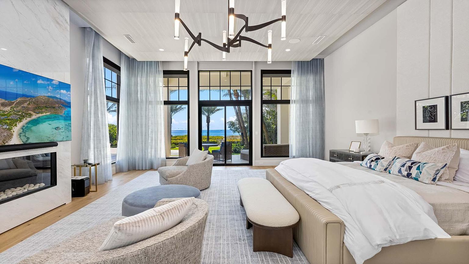 This is a luxurious primary bedroom featuring a king-size bed with a neutral-toned upholstered headboard and crisp white linens. Large windows offer stunning ocean views, complemented by sheer blue curtains that add a touch of elegance. The room is furnished with modern seating, a sleek fireplace, and stylish lighting fixtures, creating a serene and sophisticated retreat.