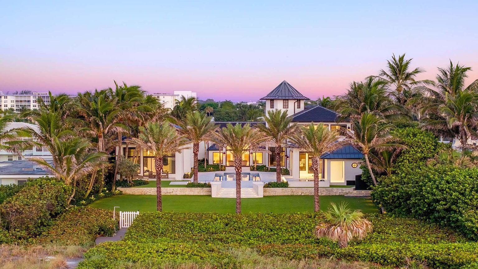 This is an exterior front view of a luxurious beachfront property. The house features a symmetrical design with a central tower element, white facade, and dark roof accents. Lush landscaping includes palm trees and manicured lawns, creating a tropical and upscale ambiance. The perspective is from a slightly elevated position, showcasing the property's grandeur and its proximity to the beach.