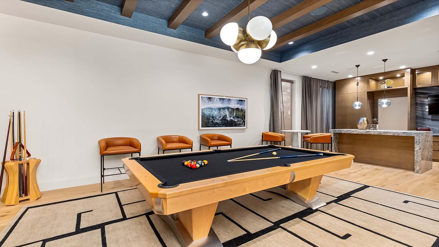 This interior shot showcases a stylish living room featuring a pool table as a focal point. The room is decorated with modern furniture, including leather chairs and a unique light fixture, complemented by a patterned rug and a piece of framed artwork. The space also includes a bar area with wooden cabinetry and marble countertops, creating an inviting and sophisticated atmosphere.