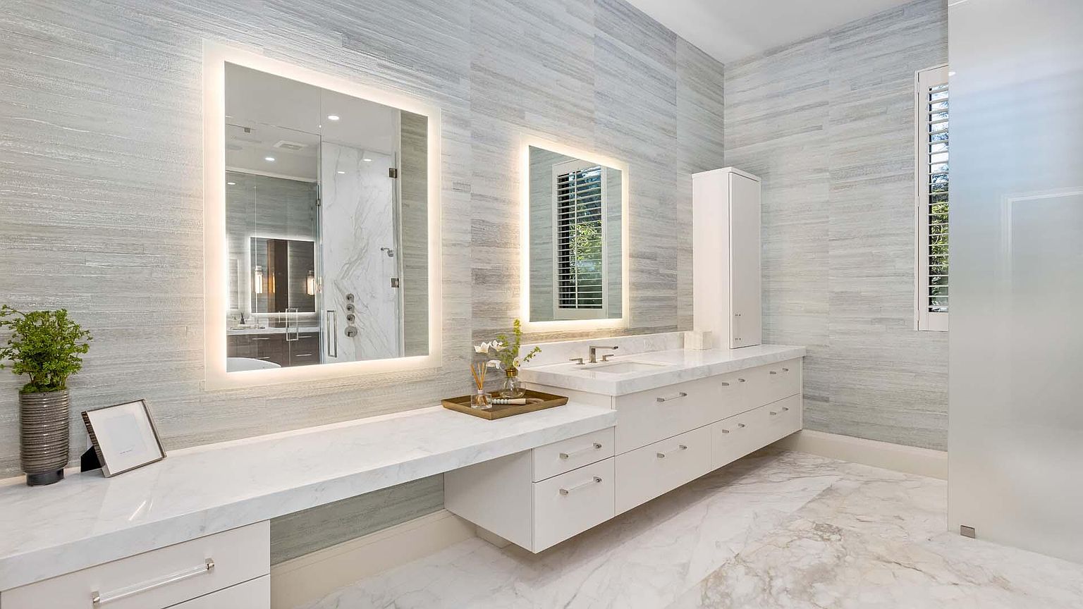This is a luxurious primary bathroom featuring a double vanity with white marble countertops and cabinetry. Two large, illuminated mirrors hang above the vanities, reflecting the spacious interior. The walls are covered in a light gray, horizontally-laid tile, and the floor is a matching marble, creating a cohesive and elegant design.