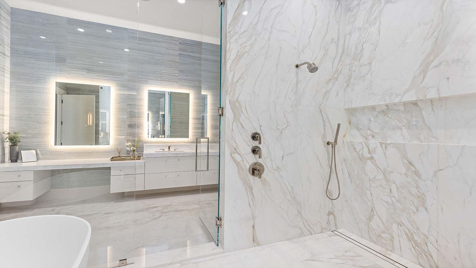 This is a luxurious primary bathroom featuring a walk-in shower with marble walls and modern fixtures. The bathroom also includes a double vanity with illuminated mirrors and sleek white cabinetry. The overall design is contemporary and elegant, creating a spa-like atmosphere.
