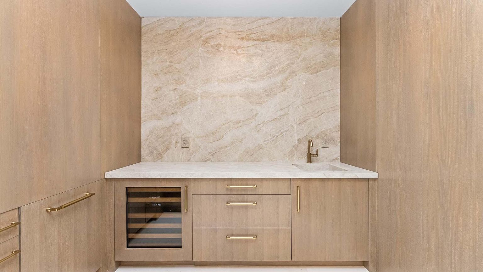 This interior shot showcases a modern wine cellar or bar area, featuring light wood cabinetry with sleek gold hardware. A built-in wine refrigerator is visible, and a marble backsplash and countertop add a touch of luxury. The overall design is clean and sophisticated, creating an inviting space for wine storage and enjoyment.