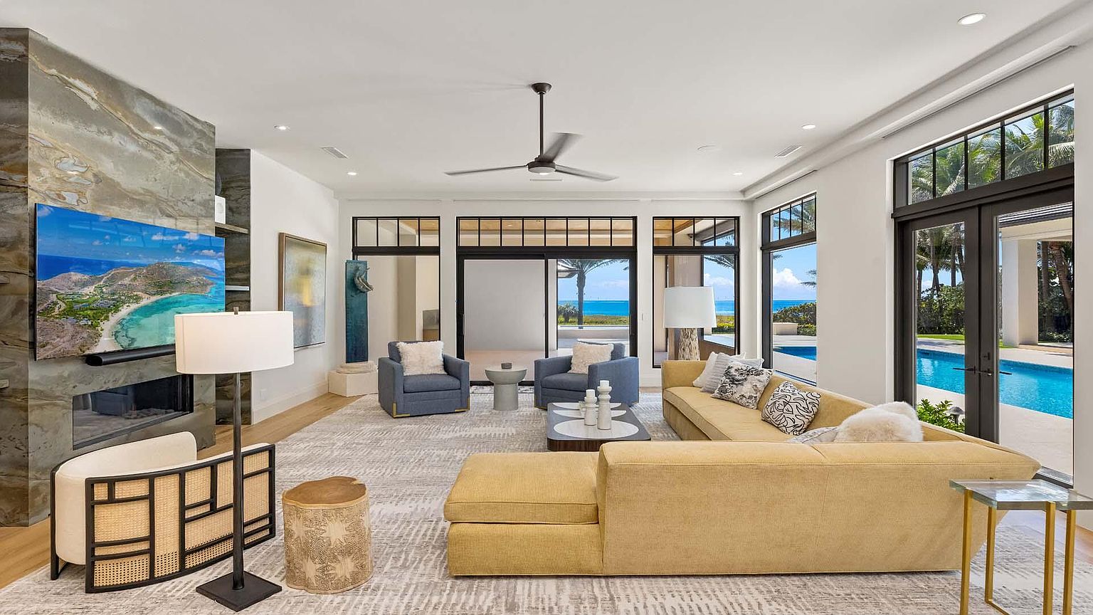 This is a bright and airy living room featuring a large sectional sofa, two armchairs, and a modern fireplace with a mounted television. The room has large windows and doors that offer views of the pool and ocean. The overall style is luxurious and contemporary, creating a comfortable and inviting space.