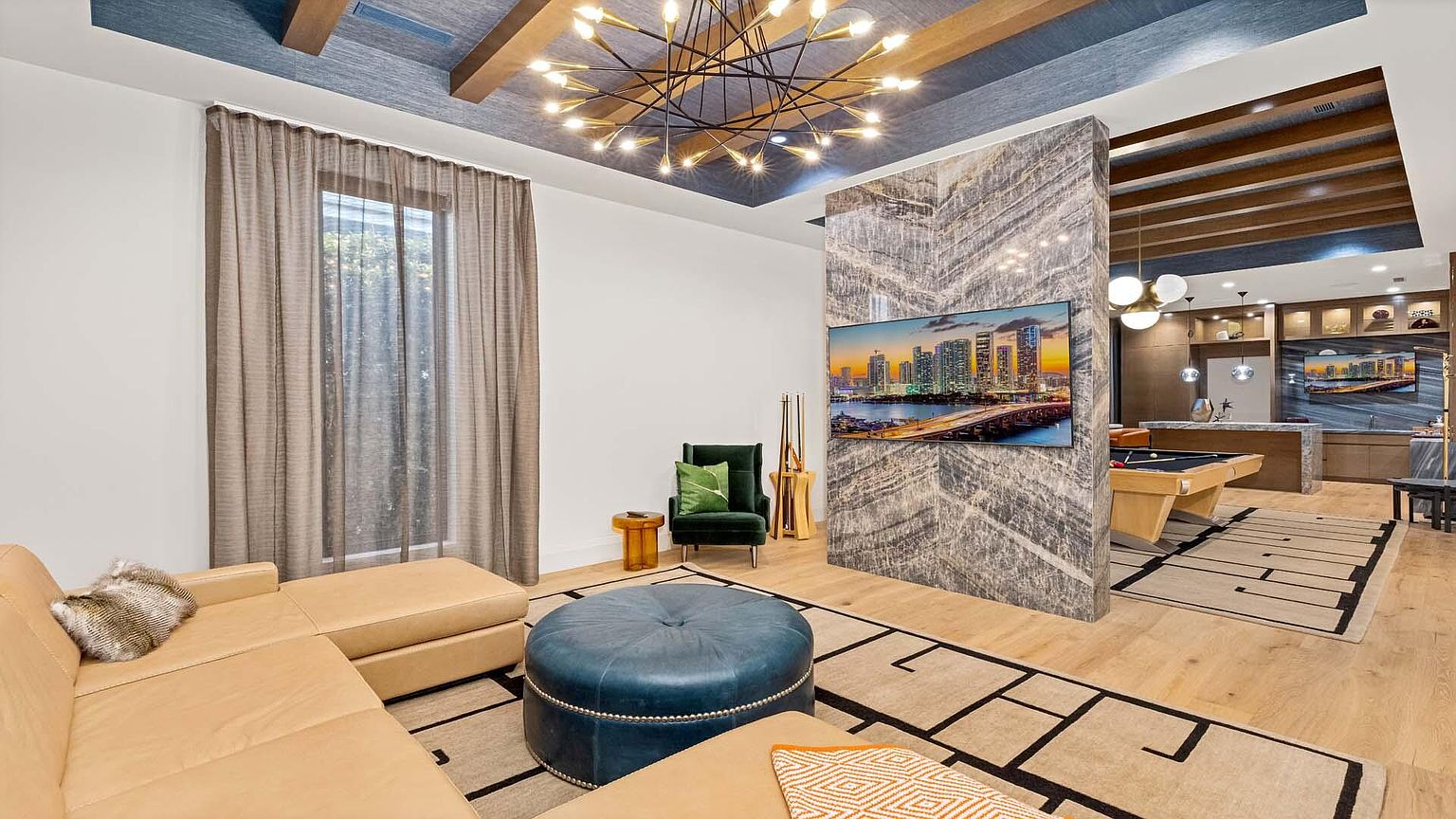 This is a stylish living room featuring a modern design with a tan sectional sofa, a blue ottoman, and a large flat-screen TV mounted on a textured wall. The room is illuminated by a unique chandelier and natural light filtering through sheer curtains. A pool table is visible in the background, suggesting a combined living and entertainment space.