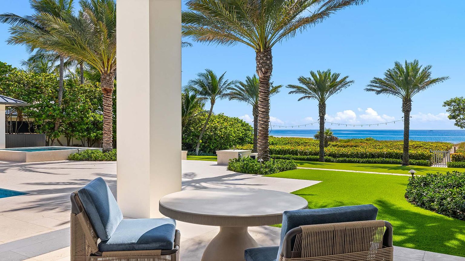 This image showcases a luxurious outdoor patio area with a round table and comfortable seating, offering a serene view of the ocean and palm trees. The well-manicured lawn and landscaping enhance the property's appeal, creating an inviting space for relaxation and entertainment. The architectural details and high-end furnishings suggest a sophisticated and upscale lifestyle.