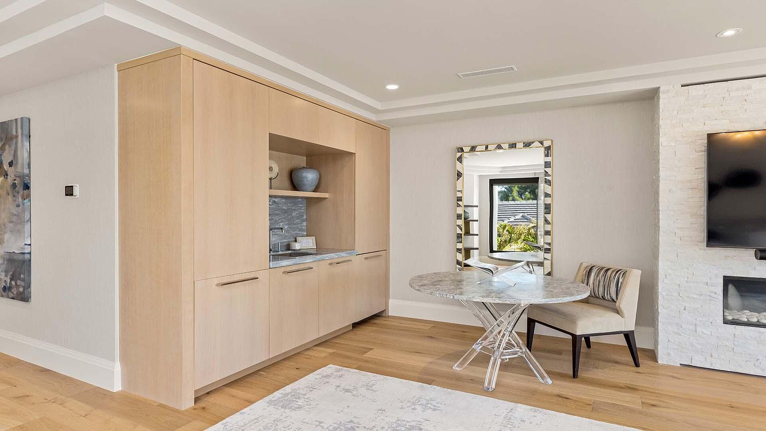 This interior shot showcases a dining area with a built-in cabinet and a round table with a marble top and clear acrylic base. A stylish chair sits beside the table, and a mirror reflects the outdoor scenery. The room features light wood flooring and a neutral color palette, creating a sophisticated and inviting atmosphere.