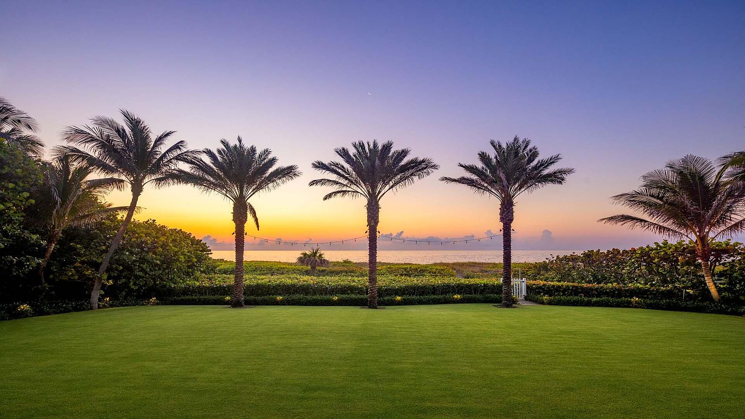 This image showcases a beautifully manicured lawn leading to a beachfront property at sunset. Palm trees line the horizon, creating a tropical and serene atmosphere. The scene is framed by lush greenery on either side, enhancing the sense of privacy and tranquility, perfect for a luxury real estate listing.
