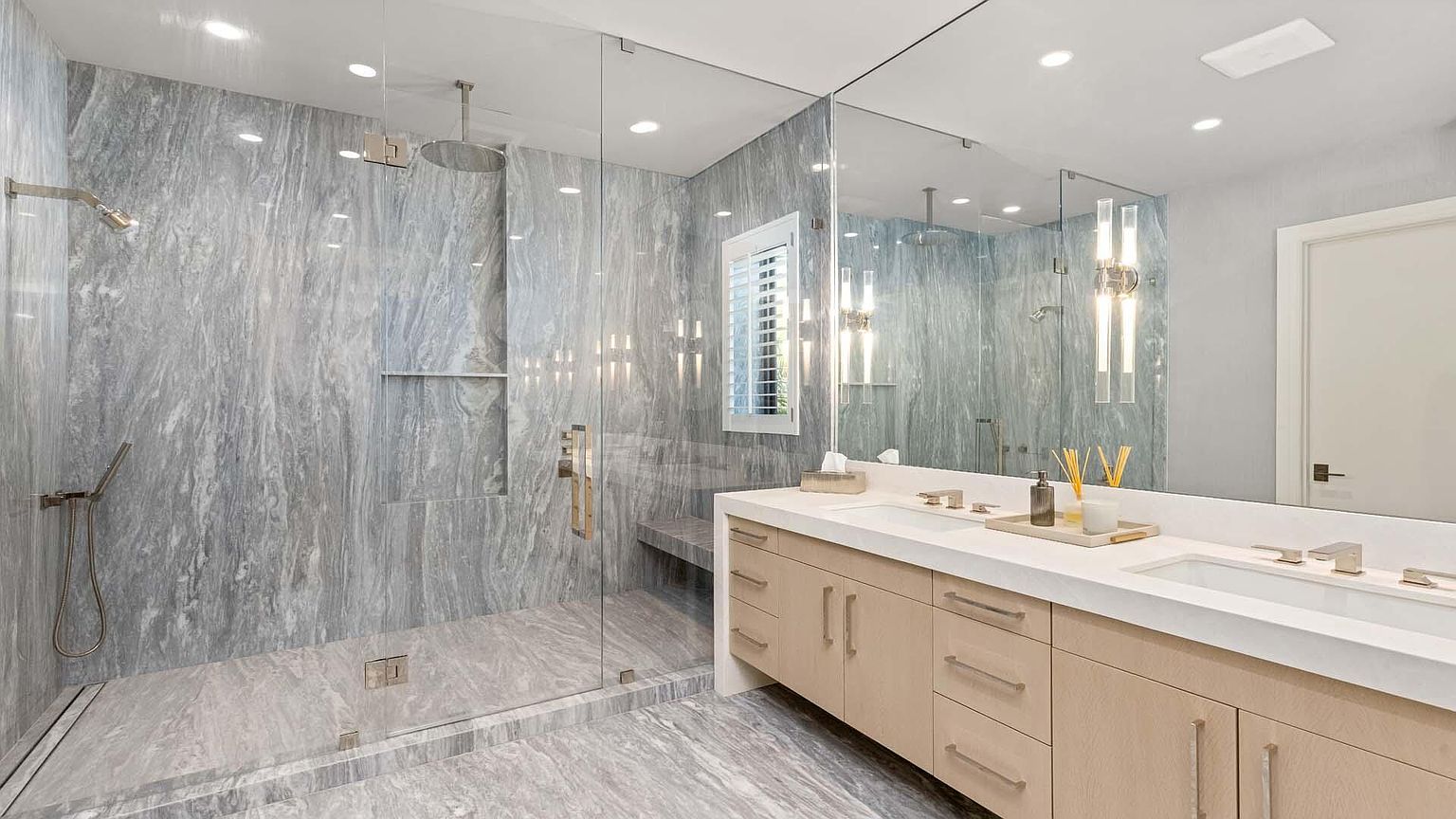 This is a luxurious primary bathroom featuring a spacious walk-in shower with gray marble-like tile and dual shower heads. The vanity area includes a double sink with modern fixtures, light wood cabinetry, and a large mirror reflecting the shower area. The overall impression is clean, modern, and spa-like.