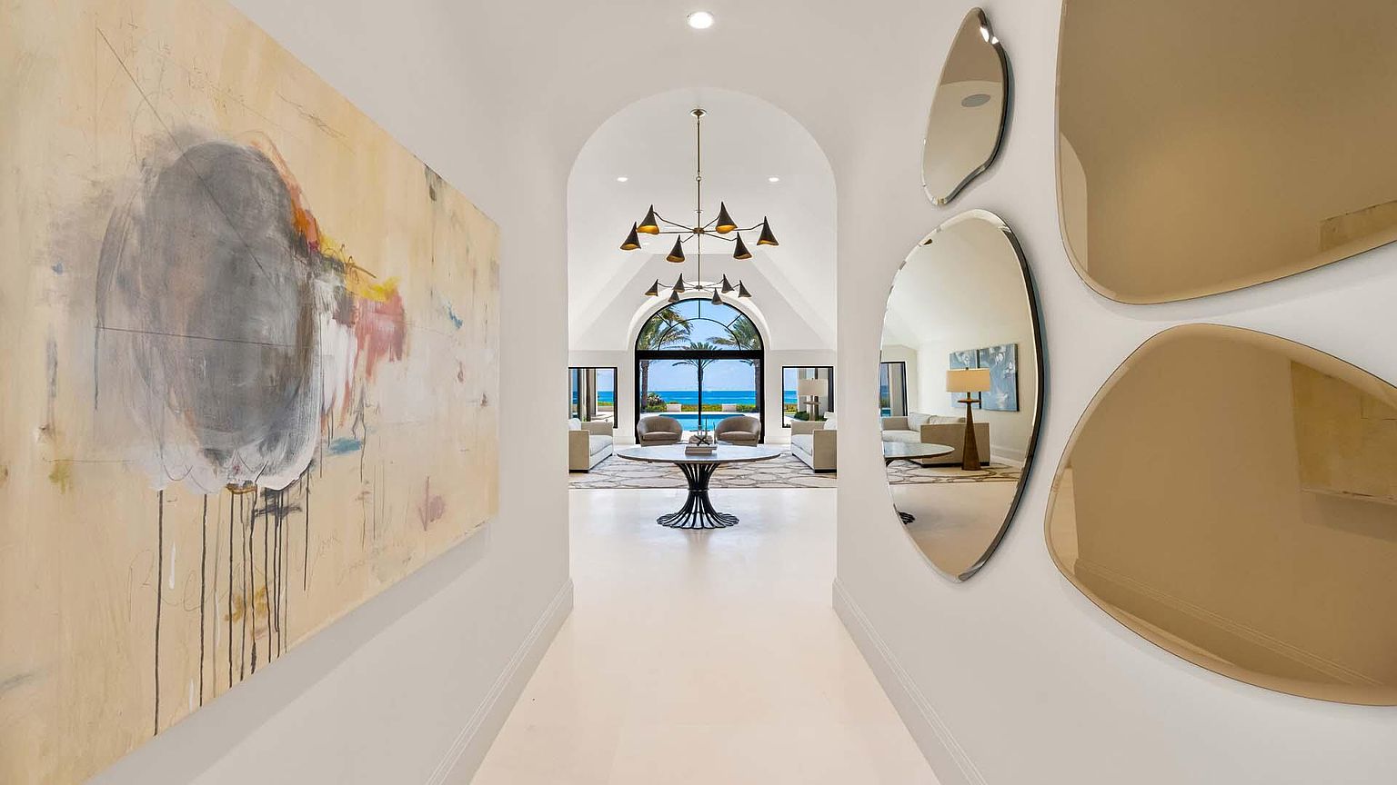 This interior shot showcases a bright hallway leading to a living area with ocean views. The hallway features a large abstract painting on one wall and decorative mirrors on the other, leading to an open living space with a modern chandelier and floor-to-ceiling windows framing a picturesque ocean scene. The design emphasizes light, space, and a seamless transition between interior and exterior.