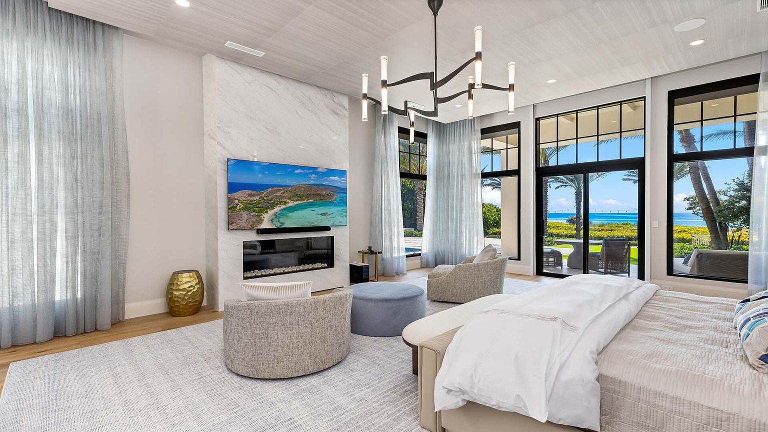 This is a luxurious primary bedroom featuring a modern fireplace with a large television above it, creating a focal point. The room is bathed in natural light from large windows offering ocean views, complemented by sheer curtains. The neutral color palette, plush seating, and contemporary chandelier contribute to a serene and upscale ambiance.