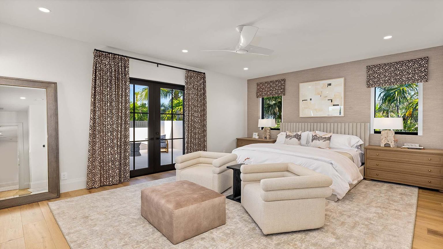 This is a primary bedroom featuring a king-size bed with a neutral-toned headboard and white bedding. The room is decorated in a contemporary style with a neutral color palette, including textured wallpaper and patterned window treatments. The room has access to an outdoor area through glass doors, and there are two armchairs and an ottoman in the foreground, creating a cozy seating area.