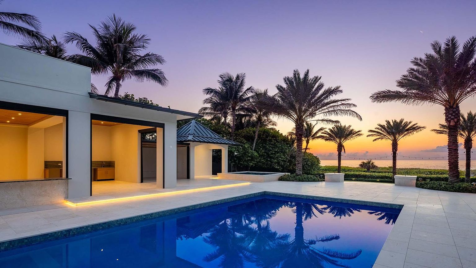 This exterior shot showcases a luxurious pool area at dusk, featuring a modern architectural style with clean lines and a serene atmosphere. The pool's reflective surface mirrors the surrounding palm trees and the colorful sunset sky, creating a tranquil oasis. The property includes an outdoor kitchen and a covered pavilion, enhancing the appeal for outdoor entertaining and relaxation.