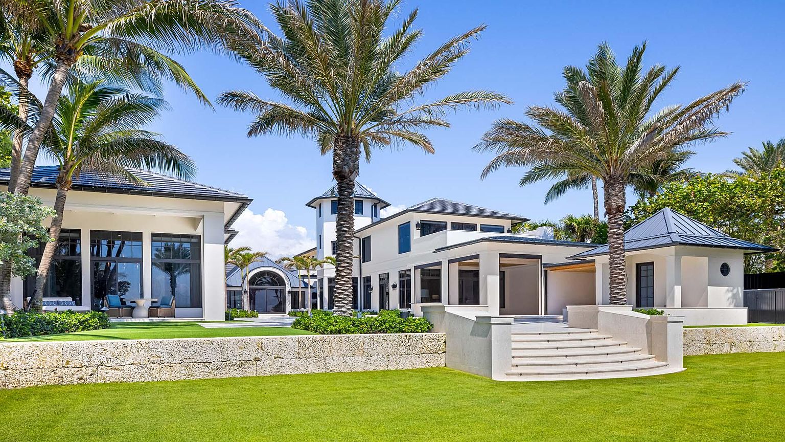 This image showcases the rear exterior of a luxurious home, featuring a multi-level design with a combination of white walls, dark-framed windows, and a dark roof. The property includes well-manicured lawns, mature palm trees, and elegant stone retaining walls, creating a sophisticated and inviting outdoor space. The perspective is a wide shot, capturing the full breadth of the property's rear facade and landscaping.