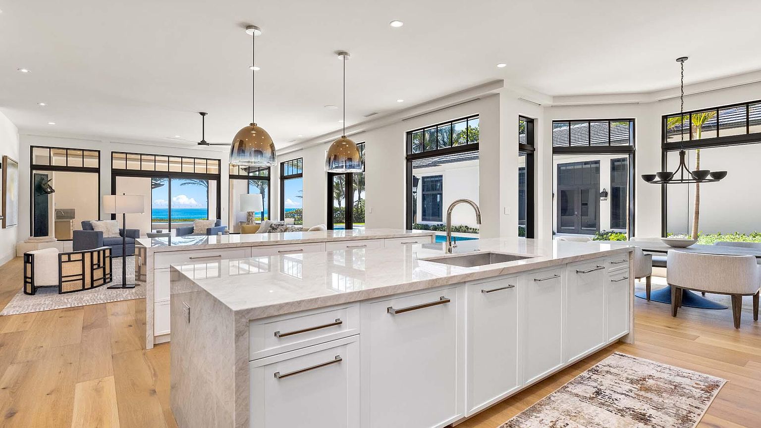 This is a bright and airy kitchen featuring a large island with a white marble countertop and white cabinetry. The kitchen seamlessly transitions into a living area with views of the ocean through large windows. Pendant lights hang above the island, adding a touch of elegance to the modern design.
